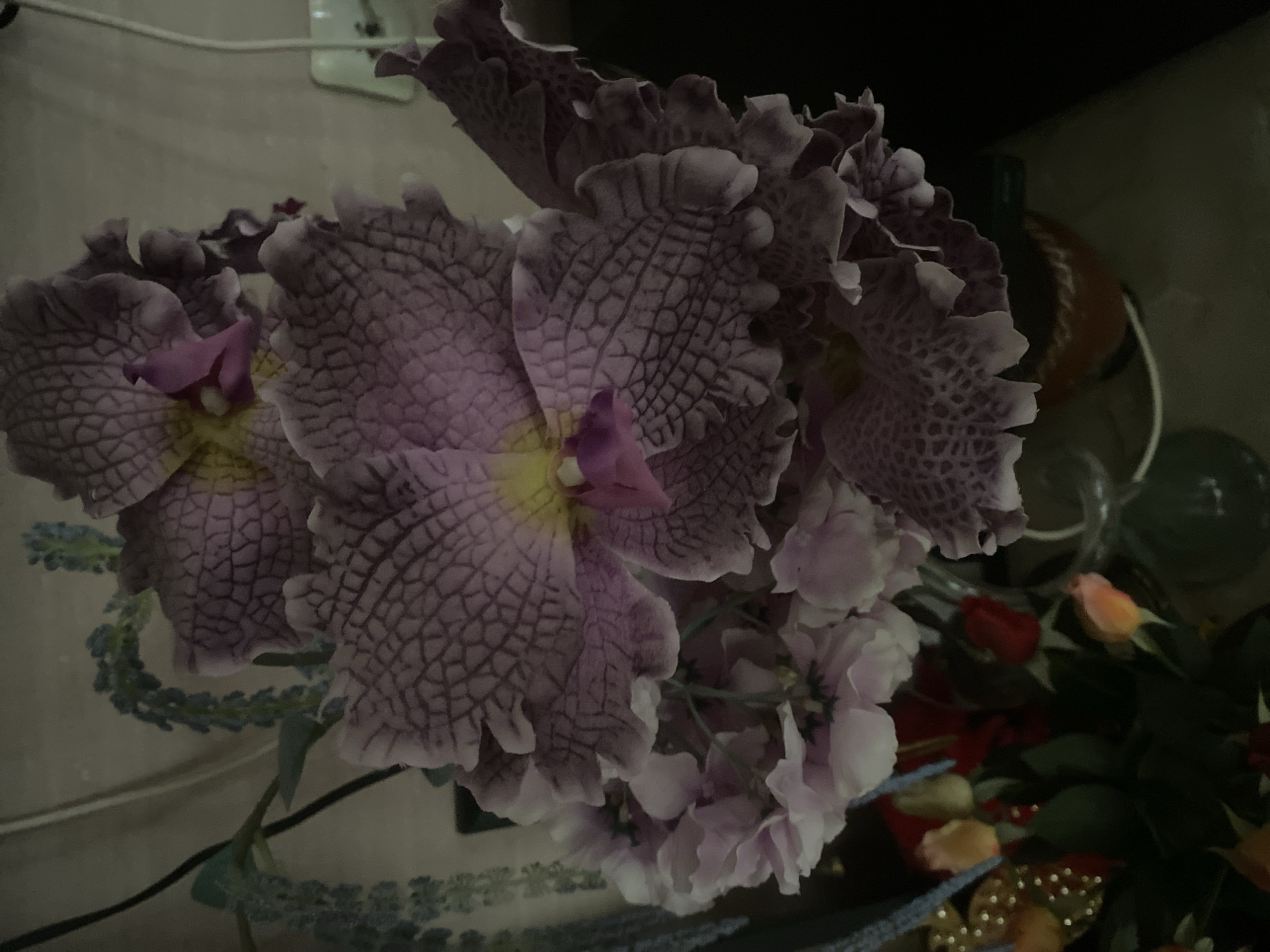 The image shows a close-up of artificial flowers, predominantly a large, intricately patterned orchid in shades of purple and pink. The orchid has ruffled petals with a textured, almost scale-like appearance. Smaller, lighter pink and white flowers are clustered beneath the orchid. A few small, colorful flower buds, appearing to be roses, are visible in the lower left portion of the frame, alongside some greenery and what looks like a decorative beaded item. The background is dimly lit and appears to be an indoor setting, with a neutral-colored wall and some electrical cords visible. There is no identifiable information about the city of Kharkiv, Ukraine, in the image itself.