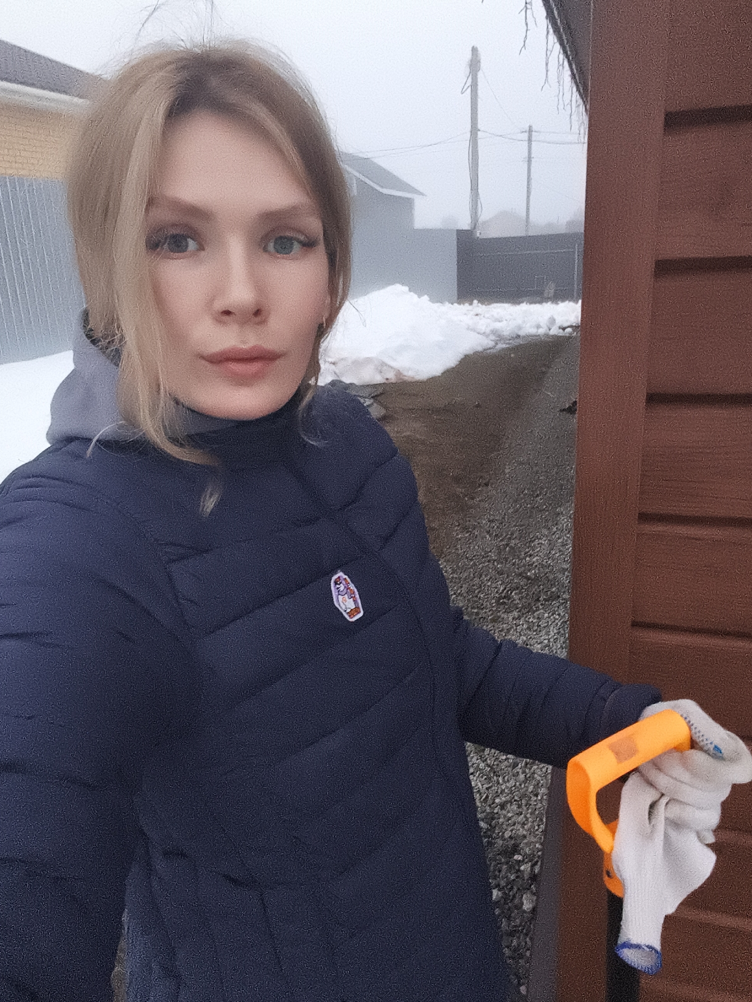 A photograph depicts a woman in what appears to be Русский Вожой, Russia. She has blonde hair and is wearing a blue puffer jacket over a gray hooded sweatshirt. She is holding a white work glove and a tool with an orange handle. Behind her, there is a path covered in gravel with patches of snow on the sides. In the background, there are houses and utility poles visible through the fog. A brown wooden structure is to her right.