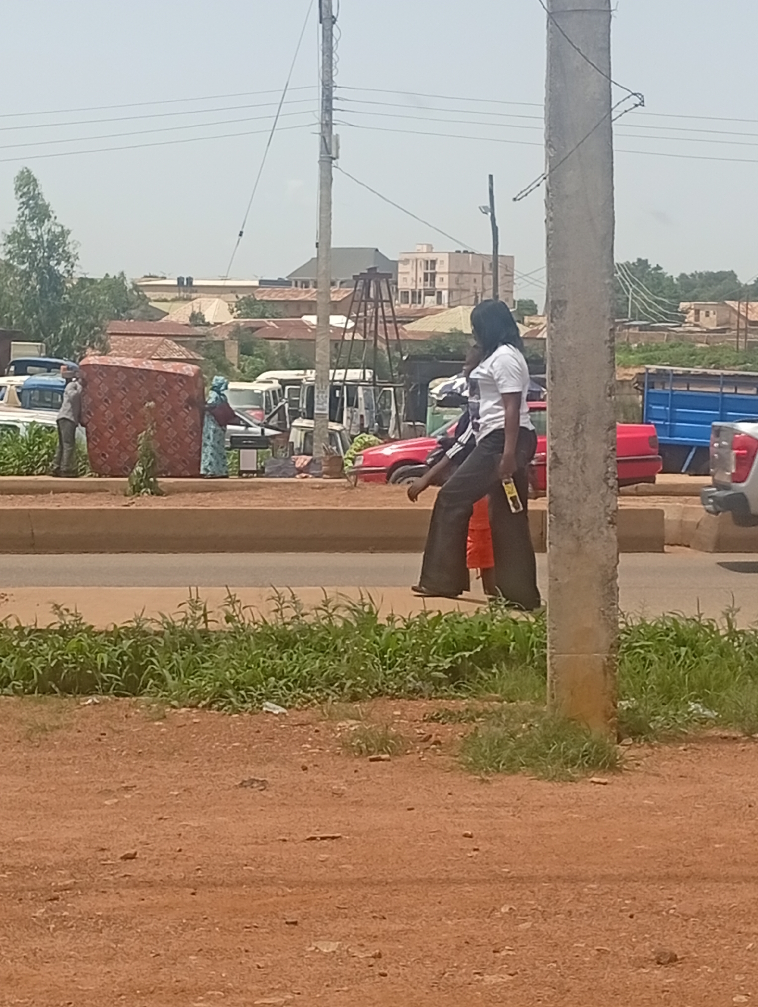 In Bukuru, Nigeria, on a clear day, a woman wearing a white t-shirt and dark, wide-legged pants walks alongside a pole. She holds a yellow object in her right hand. Behind her, a person in an orange shirt walks, followed by another individual, possibly female, wearing a blue patterned dress and holding a reddish-brown object. Further back, two men are visible. One man in a grey shirt is carrying a large, red and black patterned mattress. Adjacent to them are several vehicles, including a blue truck, a red car, and other vans and cars. The foreground is a reddish-brown dirt path with green grass and vegetation. In the background, buildings and trees are visible under a pale sky.