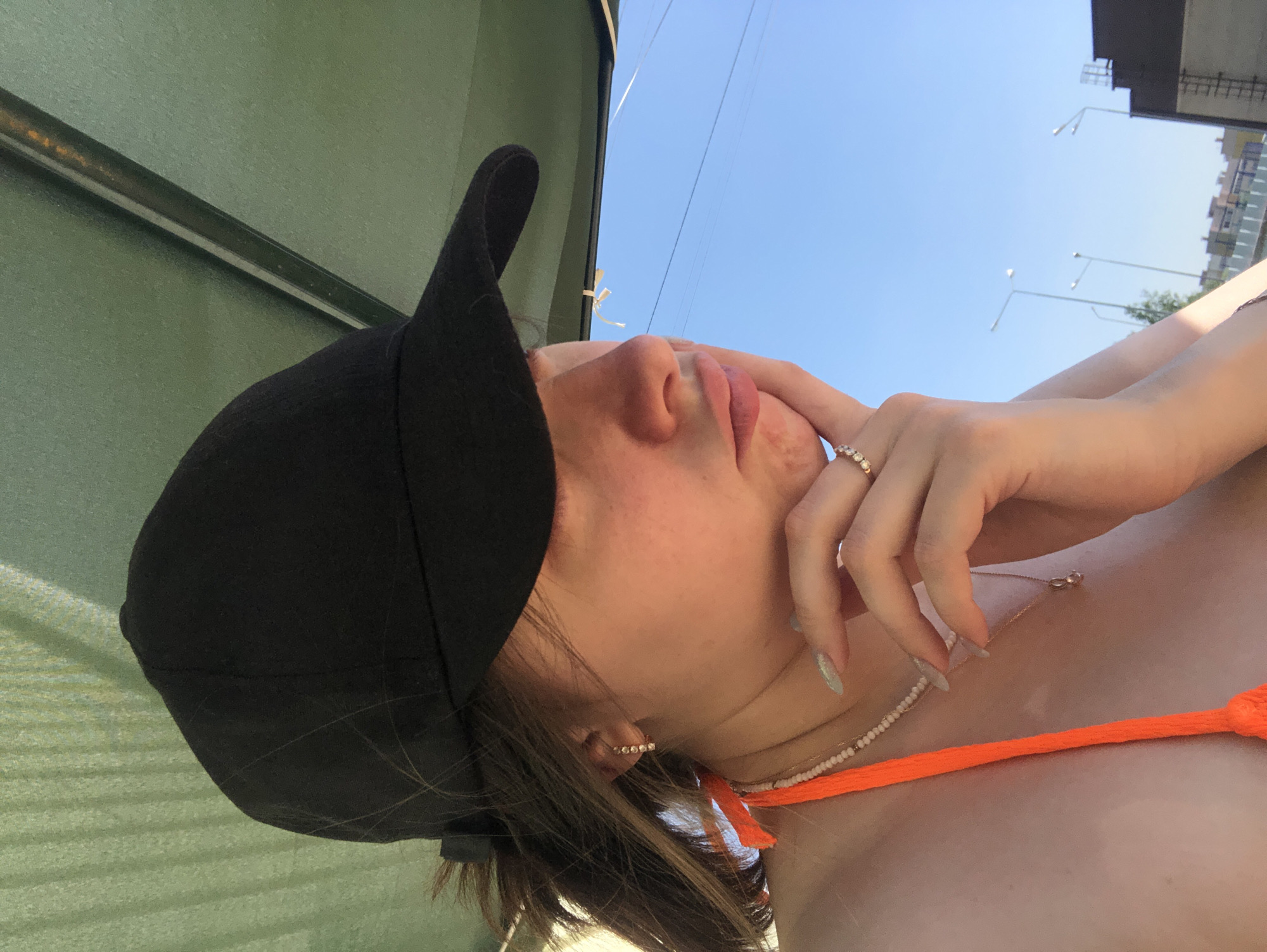 A medium-angle shot, rotated counter-clockwise, shows a light-skinned person wearing a black baseball cap and an orange bikini top. They are standing outside, with buildings and streetlights visible in the background under a clear blue sky. The person is wearing jewelry, including a ring, earrings, and necklaces. Their right hand is touching their face. The location is indicated as Noviy, Russia. There are green walls behind the person.
