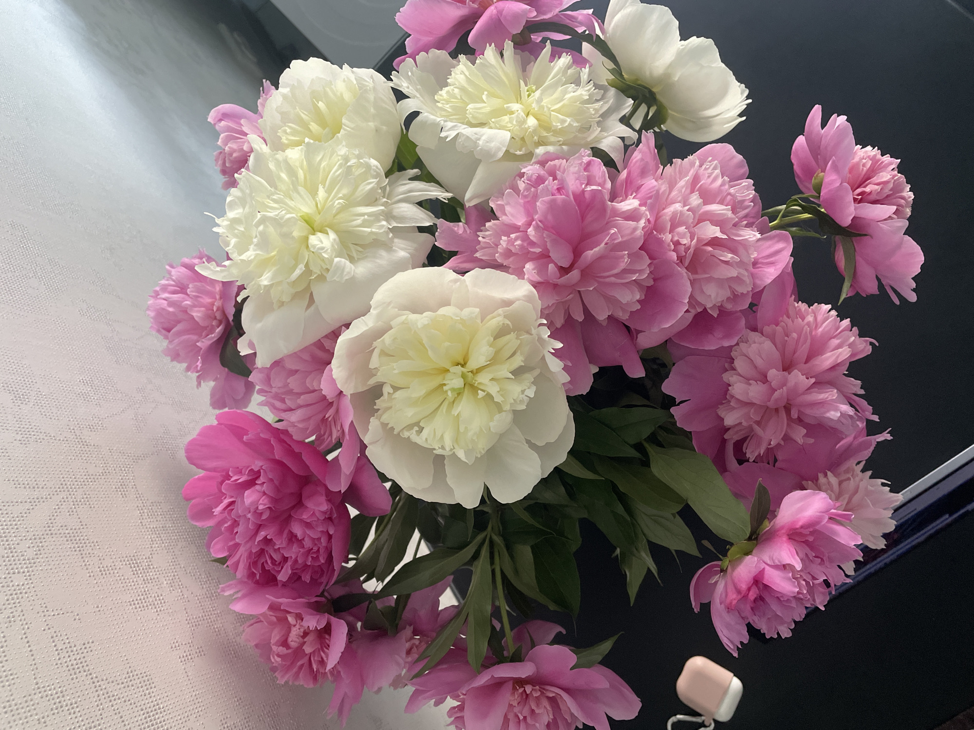 A vibrant bouquet of peonies dominates the frame, showcasing a mix of pink and white blossoms. The white peonies often feature creamy, light yellow centers, while the pink ones vary in shade. The flowers are in various stages of bloom, with many fully open and displaying abundant, ruffled petals, interspersed with dark green leaves.
The bouquet is situated on a surface that transitions across the image. On the left side, there is a light-colored, textured background, possibly a patterned tablecloth or wallpaper, with a subtle geometric design. The right side and bottom of the image feature a dark, reflective surface, likely a table, which subtly mirrors some of the flowers and light.
In the bottom right corner, resting on the dark surface, is a small, light pink or beige case for wireless earbuds, fitted with a silver-colored metal clip.
The image depicts a static scene, characteristic of a still life, and is set indoors. The lighting appears soft and natural, highlighting the textures of the petals and the surrounding surfaces. No text is visible. Based on the provided context, the location is Rozsoshentsi, Ukraine.
