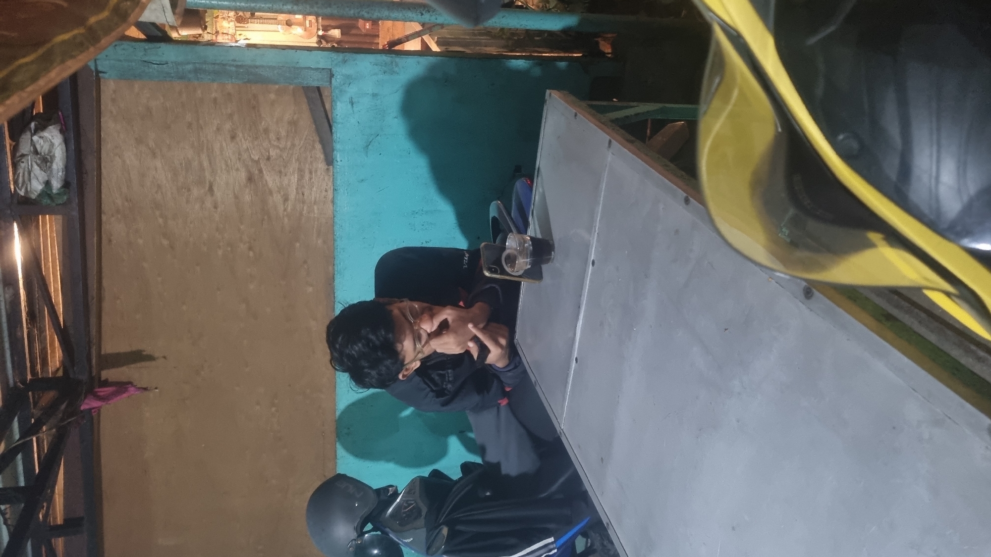 The image shows a young man with glasses sitting at a metal table in what appears to be an outdoor establishment in Kepanjen, Indonesia. He is wearing a dark jacket and is looking down at his phone, with his right hand near his face. On the table in front of him is a glass of dark liquid, possibly coffee or tea, and his smartphone. A black motorcycle helmet is visible to the left of the man, and a portion of a yellow motorcycle is in the foreground on the right. The background consists of a textured wall painted turquoise and a wooden ceiling structure. The lighting suggests it is evening or nighttime.