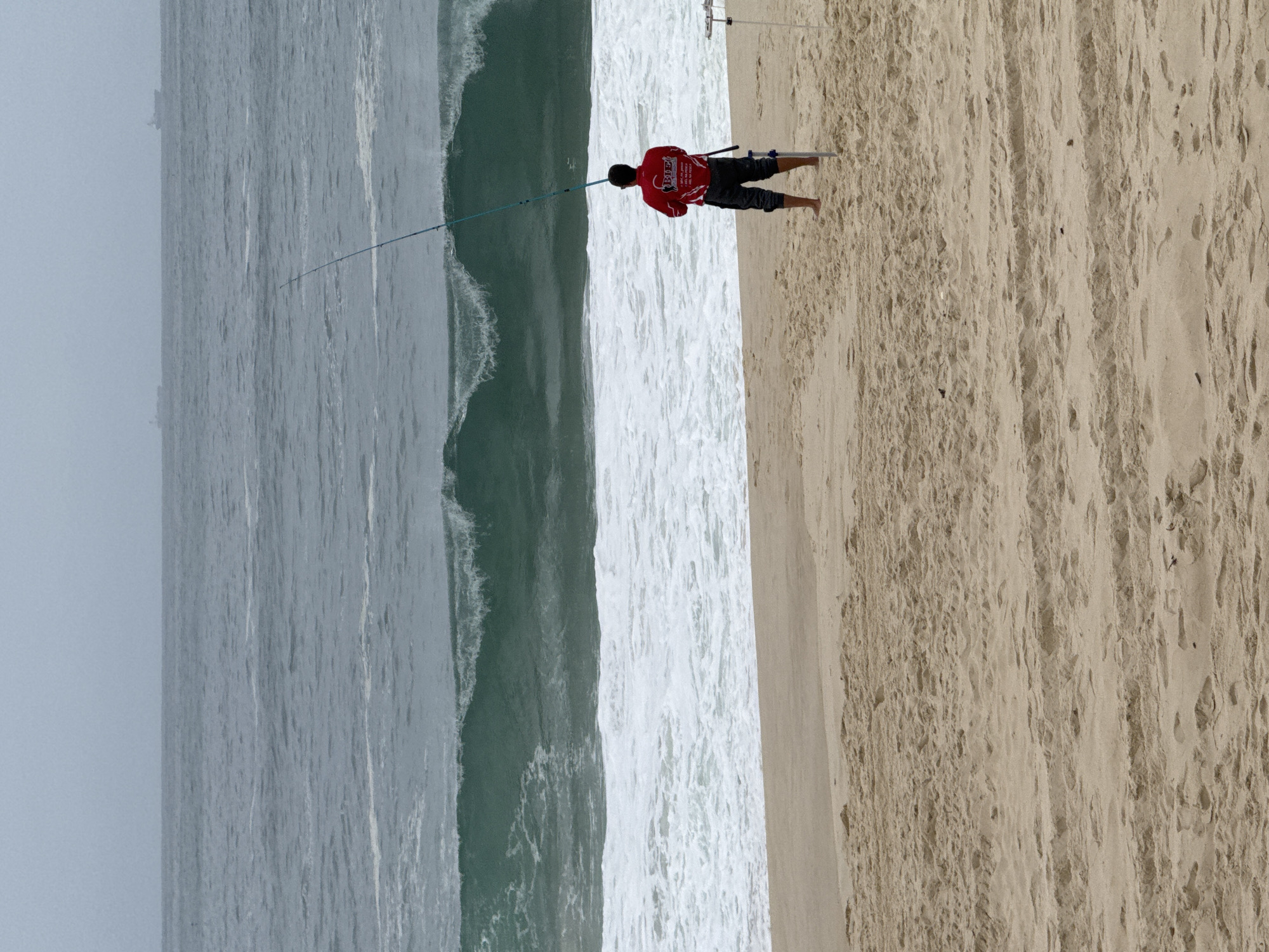 A lone fisherman stands on a sandy beach, facing a turbulent ocean. The water is a deep, dark green with white crests on the incoming waves, indicating rough conditions. Two cargo ships are faintly visible on the horizon under an overcast sky. The fisherman, wearing a red shirt with a logo and dark pants, is holding a long fishing rod, its tip angled upwards. A second, shorter fishing rod is stuck in the sand beside him. The beach is wide and appears to be made of fine, light-brown sand with some uneven textures and indentations. The scene suggests a possibly challenging day for fishing due to the waves.