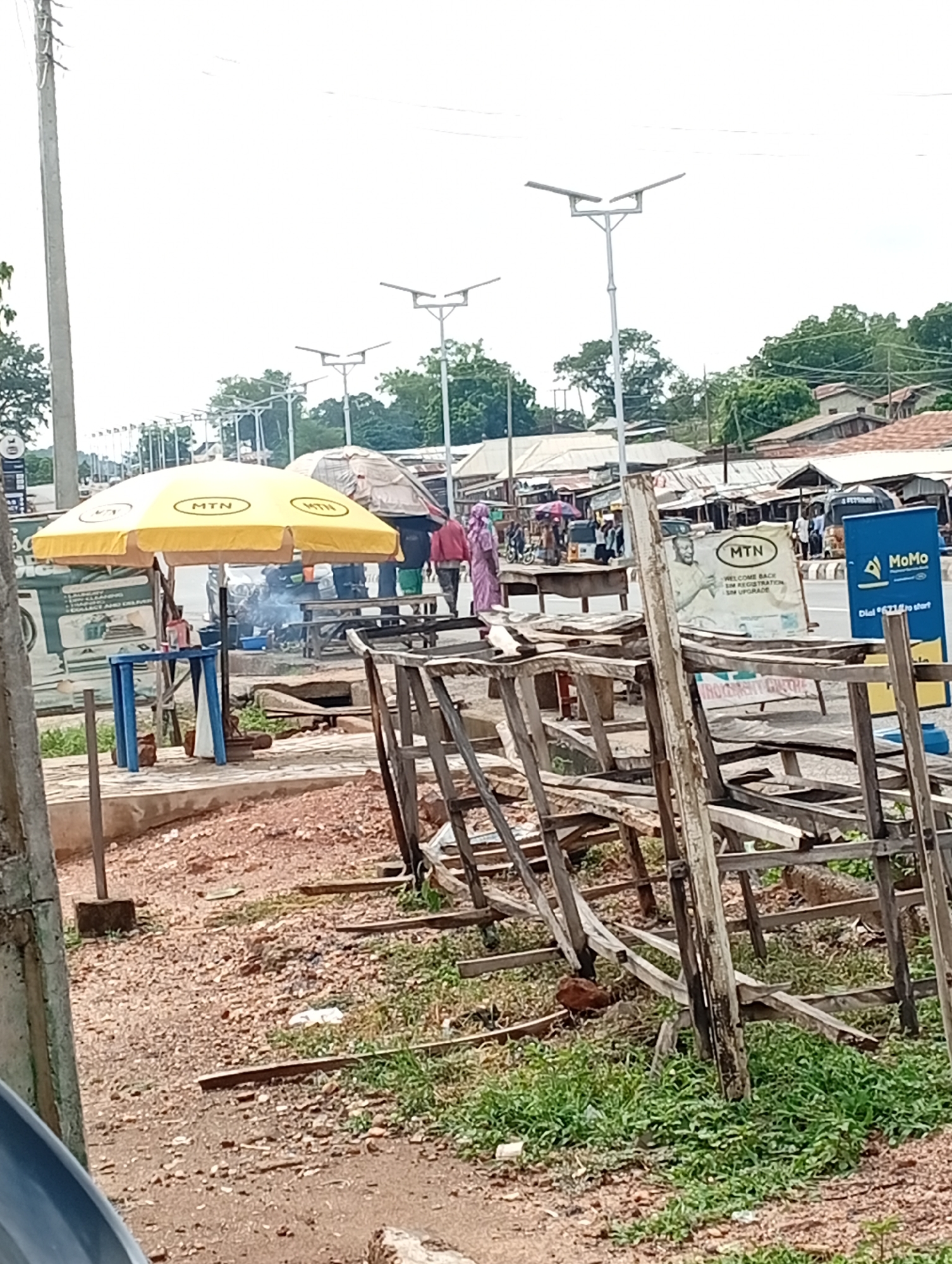 The image depicts a street scene in Adankolo, Nigeria. In the foreground, a haphazard wooden structure sits on reddish dirt with patches of grass. Behind this, there's a blue and white plastic table, likely used by a vendor. A yellow MTN-branded umbrella provides shade to an apparent outdoor cooking setup. Smoke rises from this area. Two people are walking in the background, one in pink attire. MTN and MoMo (Mobile Money) advertisements are visible, confirming a commercial presence. Solar-powered street lights line the road. Further back, buildings with corrugated metal roofs suggest a densely populated area.