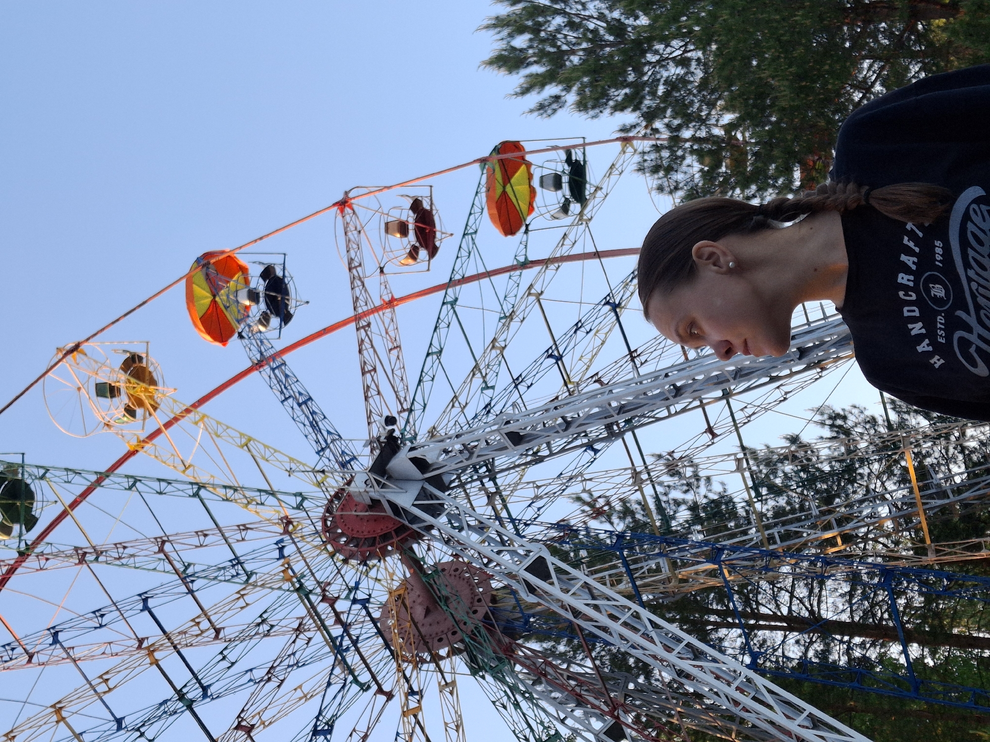 A fairground in the city of Chaikovsky, Russia, features a large Ferris wheel with multi-colored gondolas. The support structure of the wheel is a complex network of white, blue, yellow, and red metal beams. A person with a dark braid is standing nearby, wearing a dark shirt with the words "HANDCRAFT Heritage ESTD 1985" visible. Trees surround the attraction, and the sky is a clear, pale blue.