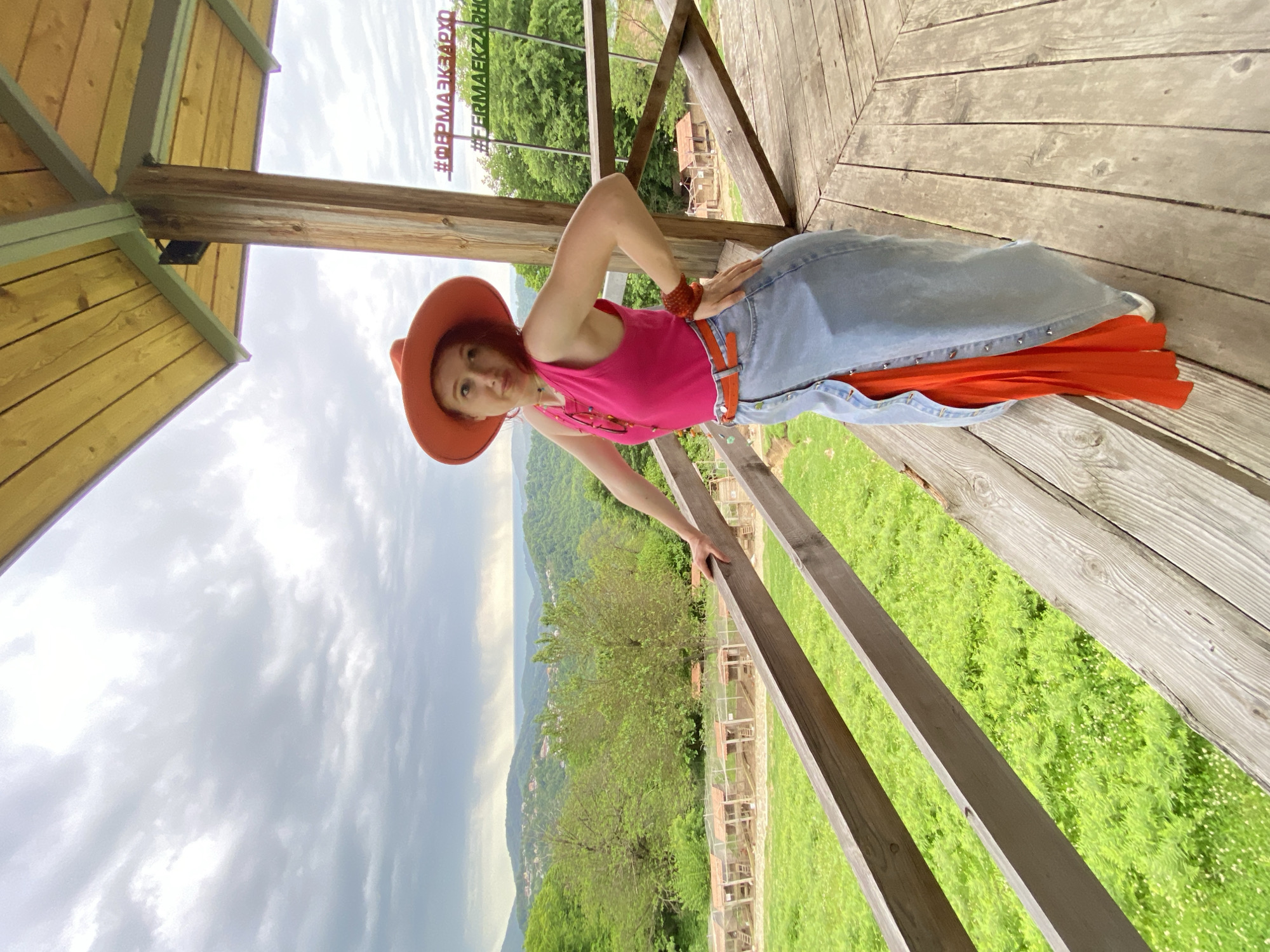 A woman with red hair wearing an orange hat, pink tank top, jeans skirt, and orange belt is posing on a wooden platform with a railing. She is standing on the platform and leaning against the railing with her right hand. She is looking at the camera with a slight smirk. In the background, there is a lush green landscape, including trees, hills, and a cloudy sky. The hashtags #ФЕРМАЭКЗАРХО appear in the upper left. The location is likely in Прогресс, Russia, near a tourist destination or attraction. The woman is possibly engaging in a photoshoot or documenting her visit.