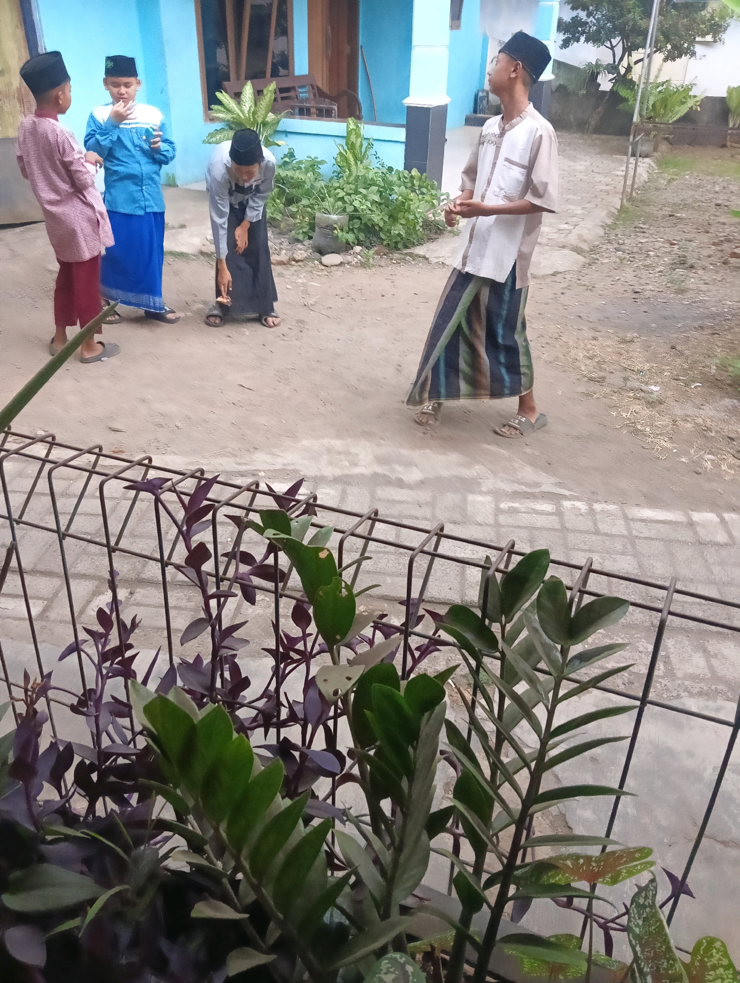 Four individuals, three of whom appear to be young males and one slightly older male, are standing in a dusty, outdoor area in front of a light blue building. The three younger males are dressed in traditional Islamic attire, including peci hats and sarongs, with one wearing a light pink and red patterned tunic, another in a blue and white striped tunic, and the third in a grey tunic. The older male is wearing a light-colored tunic and a patterned sarong, with his back to the camera, looking to the right. The ground is dirt and gravel, with some patches of vegetation and a black metal fence with plants in the foreground. The overall scene suggests a casual gathering or interaction in a residential or communal outdoor space in Sokaraja, Indonesia.