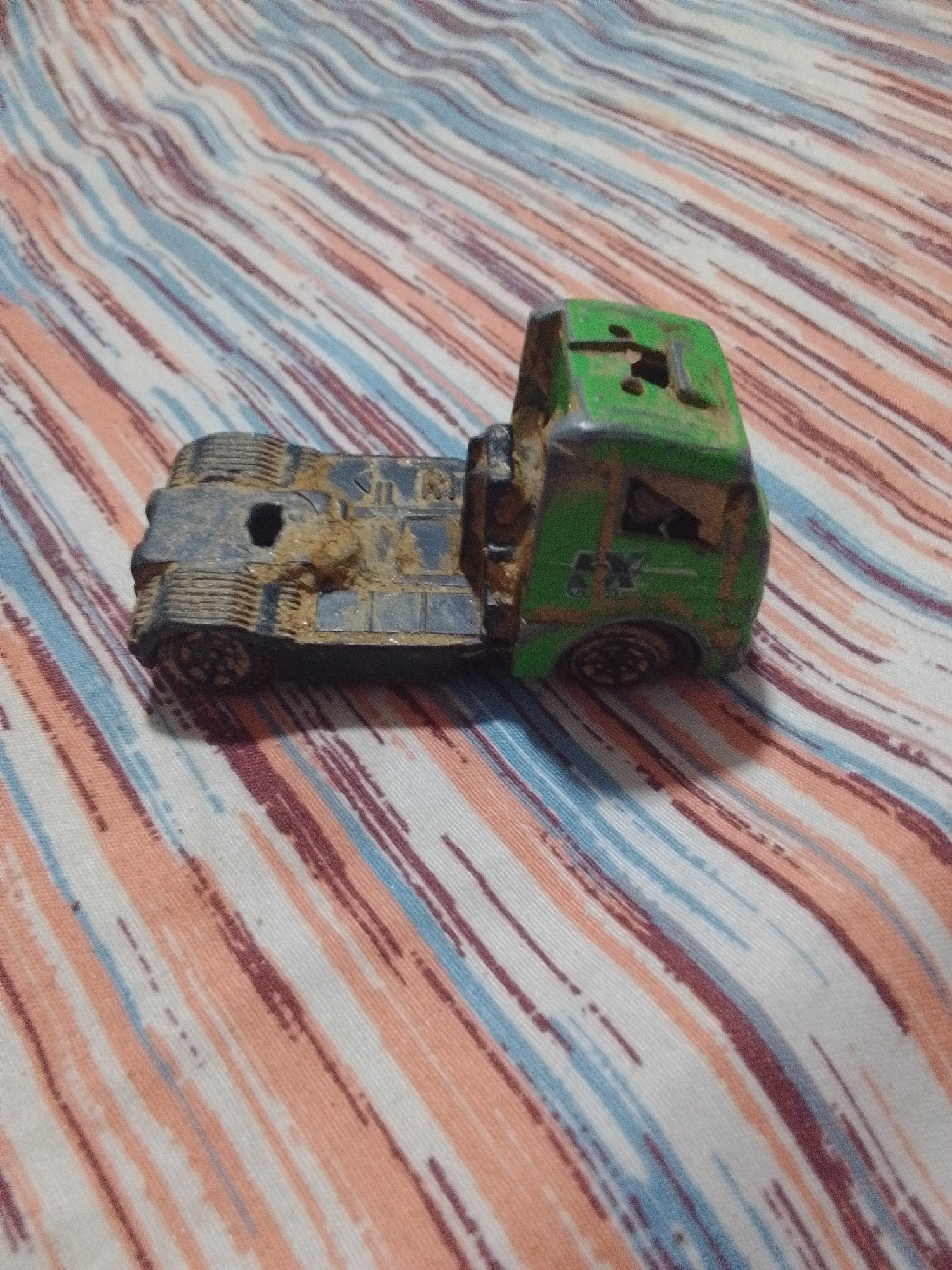 A dirty, green and black toy truck is lying on its side on a striped fabric surface. The fabric has alternating diagonal stripes of white, peach, blue, and dark red. The toy truck appears to be made of plastic and metal. The front part of the truck, including the cabin, is green, while the rear part, including the wheels and chassis, is black. The truck is covered in what appears to be dirt or mud, with some of the dirt obscuring details of the toy. The overall condition of the toy suggests it has been played with or exposed to the elements. There is no indication of any specific location within La Tagua, Venezuela.