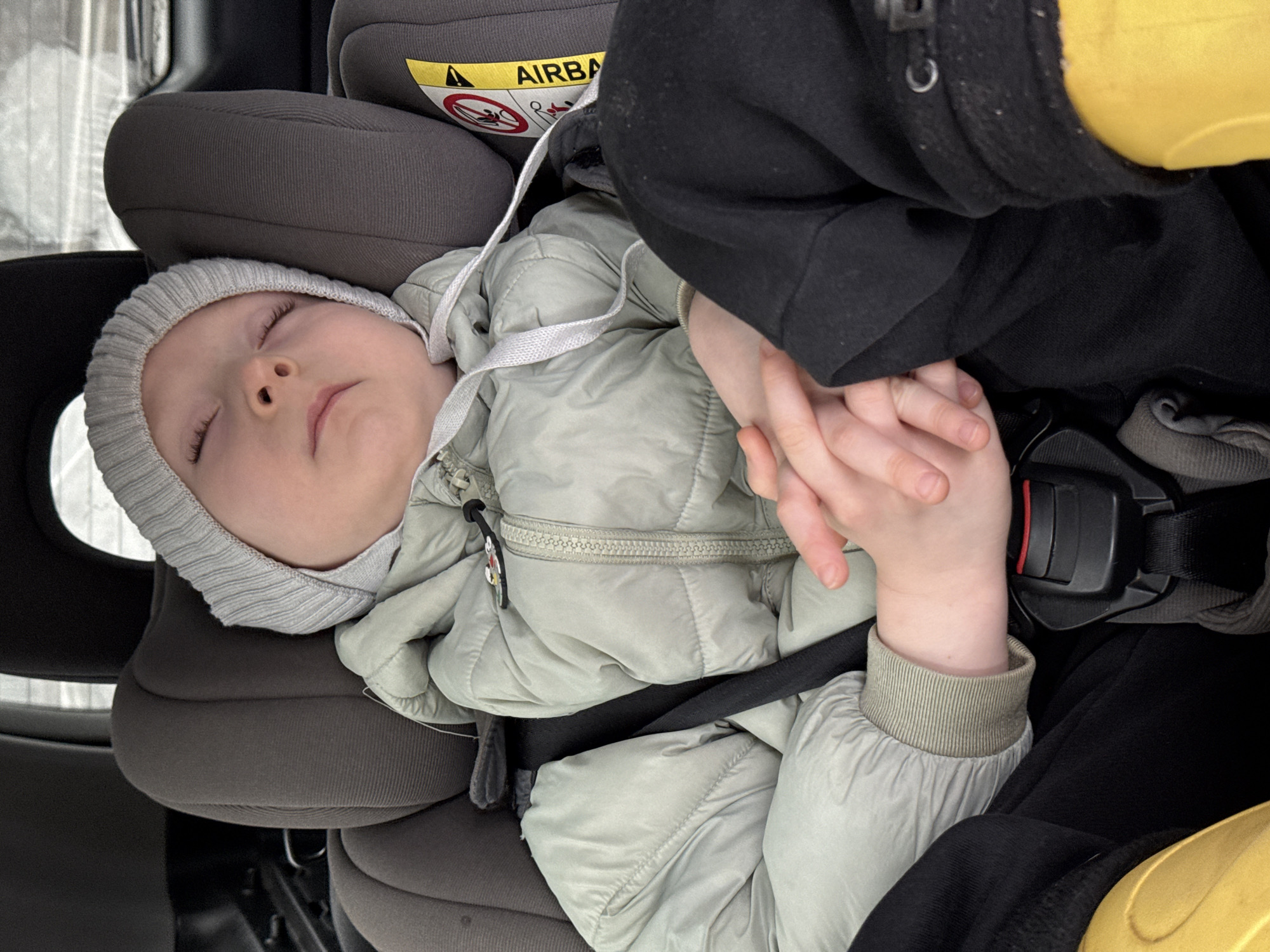The media file captures a peaceful scene of a baby sleeping soundly in a car seat, likely within a vehicle traveling in Pavlovskii, Russia. The infant is dressed warmly in a light sage green puffy jacket and a light grey ribbed beanie, indicative of cold weather. A blurry, light-colored landscape, possibly snowy, is visible through the car window, reinforcing the winter setting.
The baby is secured in a dark grey car seat with a prominent headrest. An adult's hand, partially visible on the right, gently holds the baby's hands, suggesting comfort and reassurance during the journey. The car seat's safety features are visible, including a black harness strap across the baby's lap. A yellow warning label on the car seat headrest clearly shows the text "AIRBAG" and symbols advising against placing a child seat in front of an active airbag. A small, colorful charm, possibly a cartoon character, is attached to the zipper of the baby's jacket. The baby's closed eyes and relaxed posture convey a sense of deep sleep and contentment.