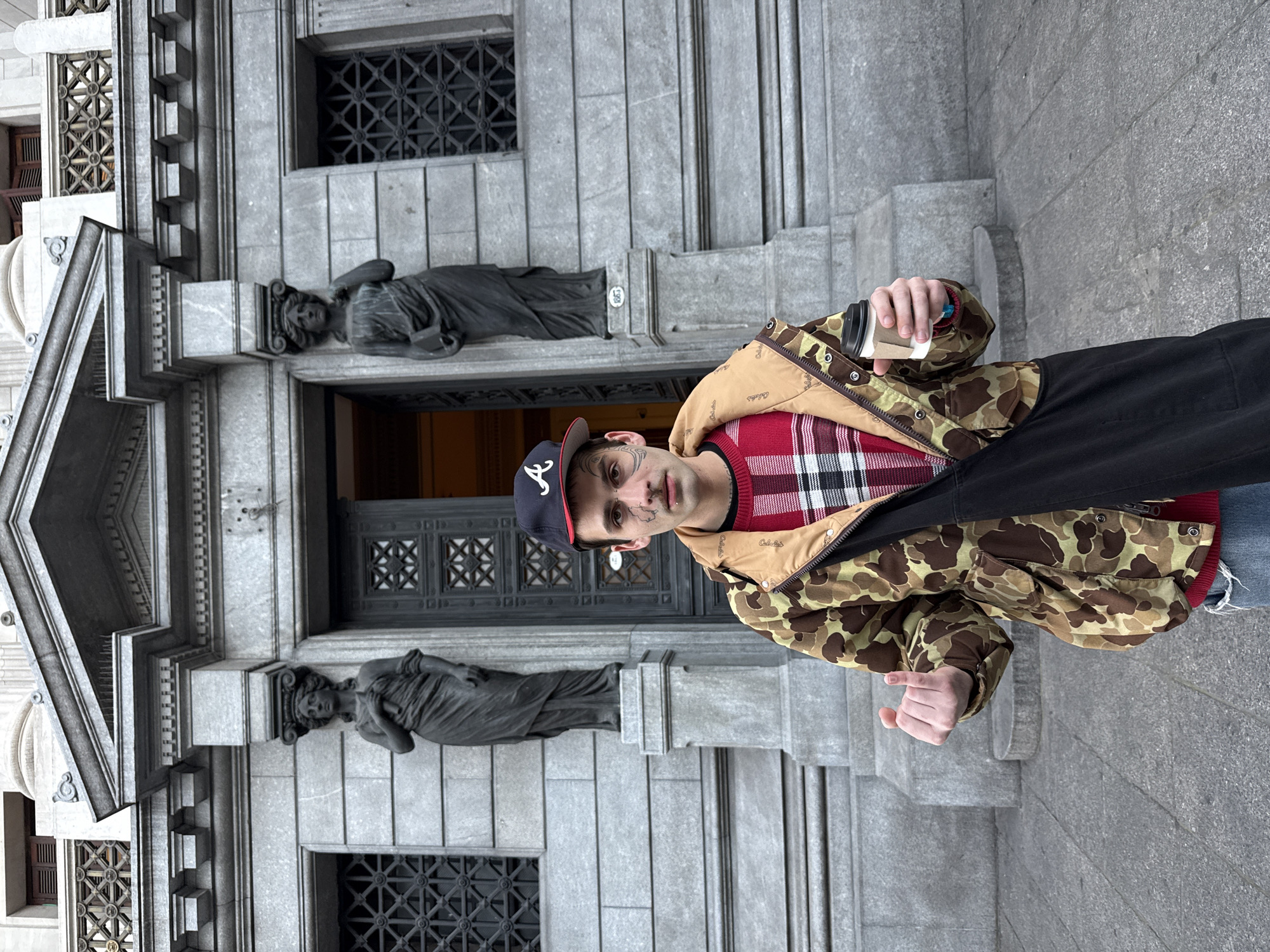 The image shows a man standing in front of the Recoleta Cemetery in Buenos Aires, Argentina. The man is wearing a baseball cap, a red plaid sweater, and a camouflage jacket. He is holding a cup in his right hand and is standing on the steps leading to the cemetery entrance. Two reclining statues flank the doorway. The cemetery is made of grey stone and features a large door.