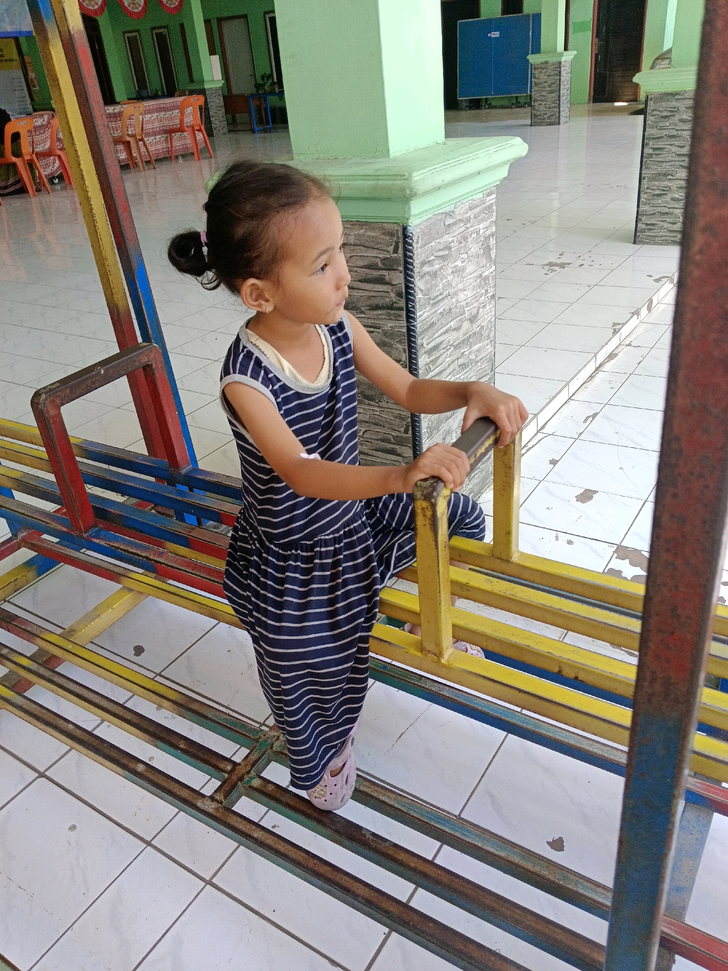 A young girl with dark hair pulled into a bun and wearing a blue and white striped dress stands on a multi-colored metal playground structure. She is looking to her right. The playground is located in a tiled outdoor area, likely part of a school or community center. In the background, there are green walls, stone pillars, and some chairs. The scene appears to be in Krembangan, Indonesia.