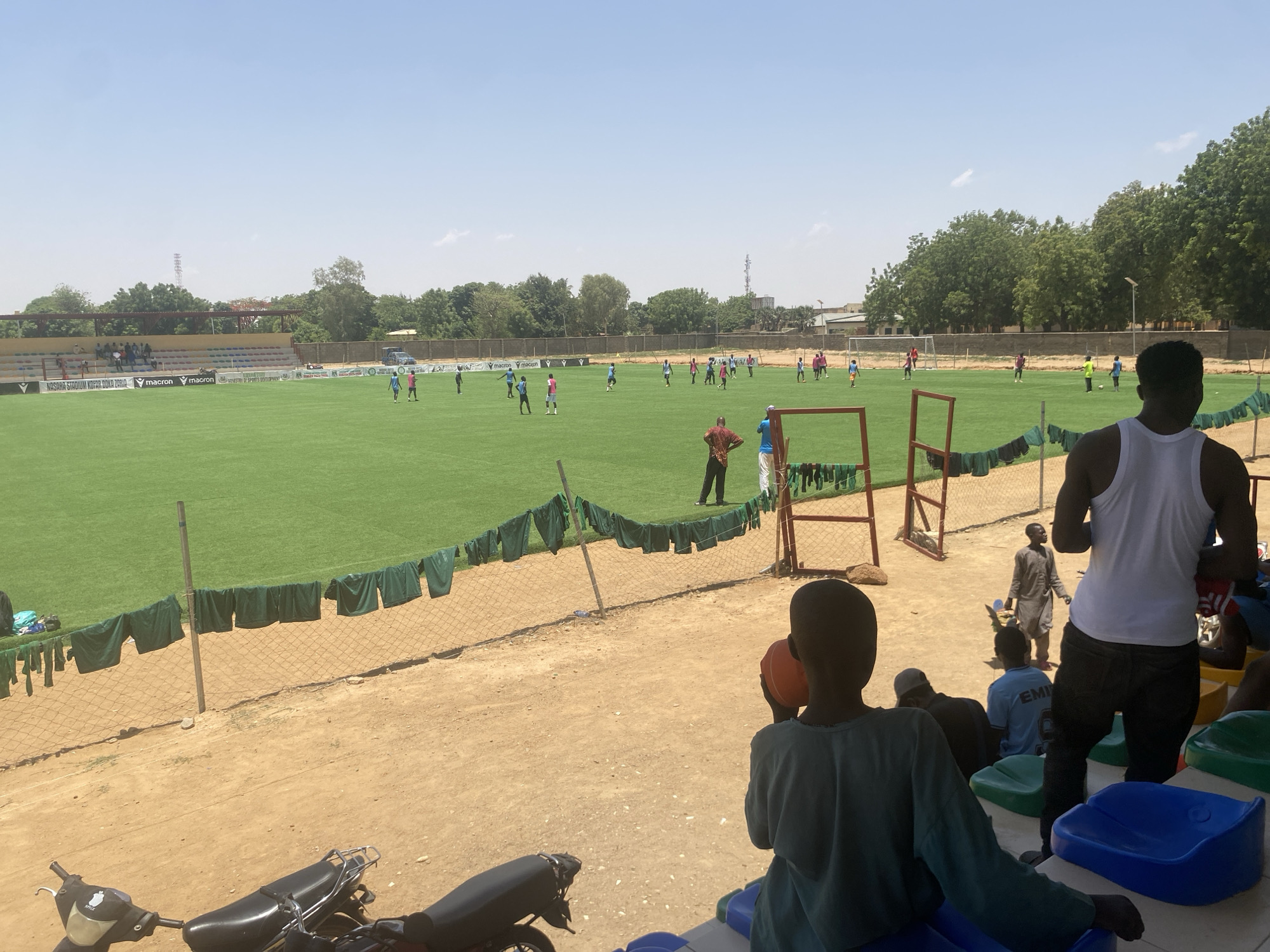 The image depicts a soccer game in progress at a stadium in Tudun Wada, Nigeria. Several players are on the green field. Spectators are in the foreground, some seated in colored plastic seats, others standing. A motorbike is parked near the lower-left corner. Dark green shirts are hanging on the wire fence. One man in a red shirt is walking on the field near a clothes-drying rack. A person in a white tank top is standing among the spectators. The sky is clear.