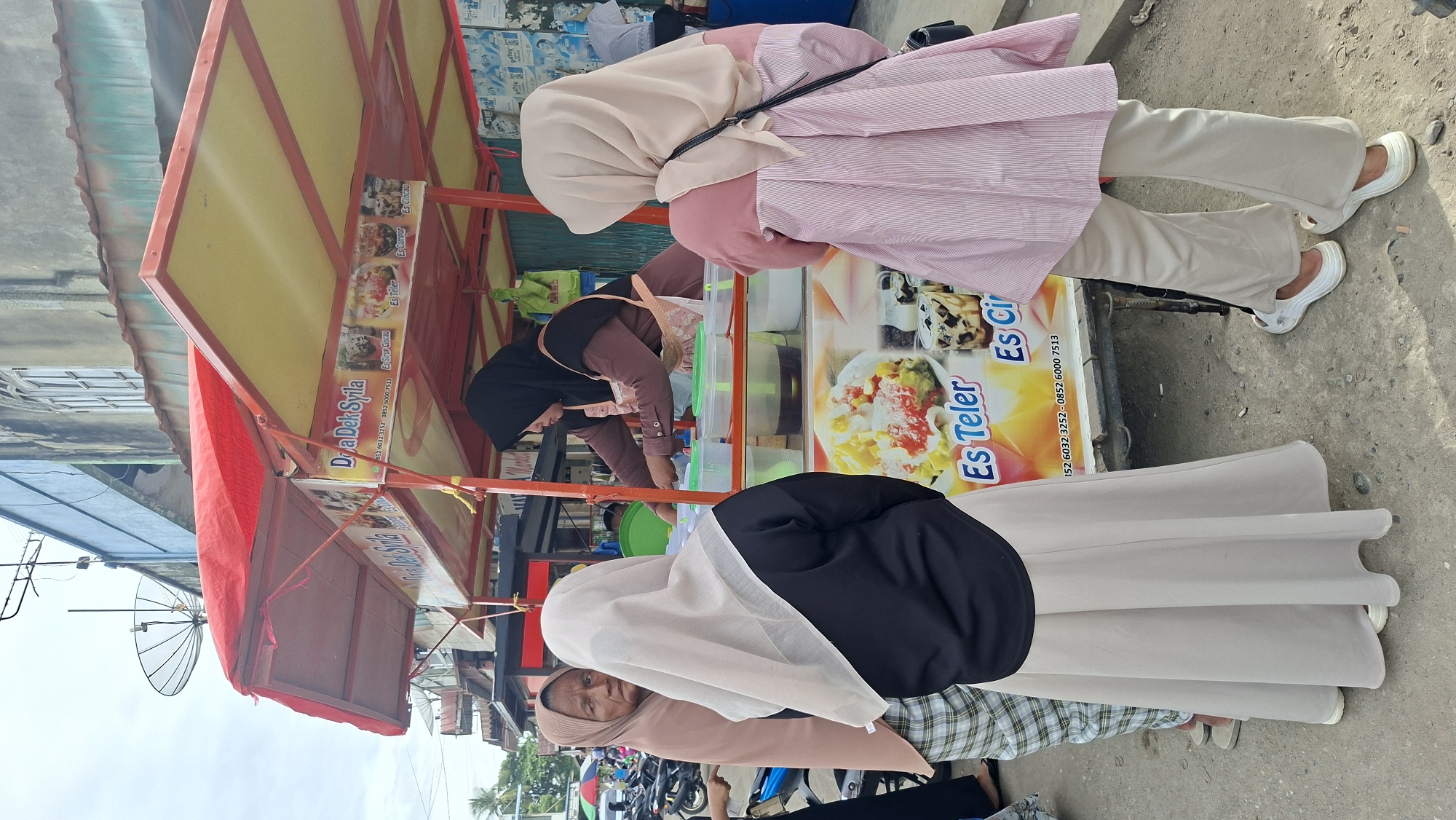 In Keudegeudong, Indonesia, a street-side food stall is busy serving customers. The stall is adorned with a red and yellow awning and displays colorful advertisements for various Indonesian desserts, including "Es Teler," "Es Campur," and "Es Sirup Cincau." The text on the stall, "Doa Dek Syila," likely indicates the name of the business.
Several individuals are visible, primarily women wearing hijabs. One woman, in a pink striped top and light-colored pants, is standing near the stall. Another woman, wearing a black hijab and a pink top, is actively preparing a drink behind the counter, reaching into containers filled with ingredients. A third person, in a beige hijab and a patterned skirt, is seated closer to the foreground. The scene appears to be set during the daytime, under what seems to be bright or sunny weather, given the lighting. The background suggests an outdoor, urban environment with buildings and parked motorcycles. The overall impression is of a lively, everyday commercial activity at a local food vendor.