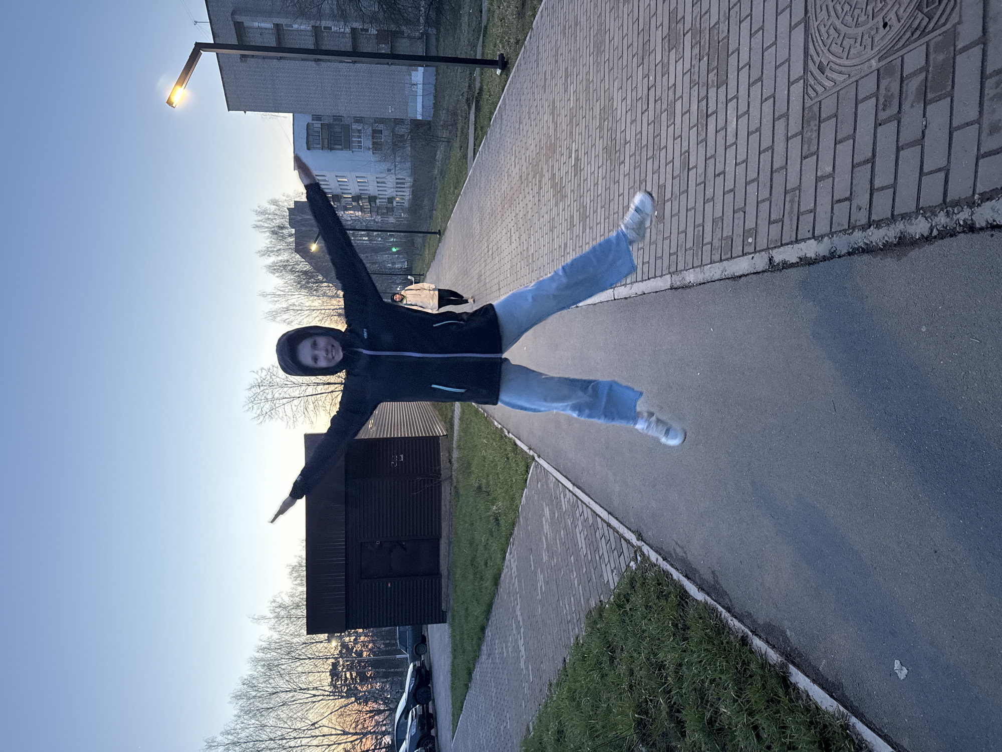 The image, when correctly oriented, captures a dynamic scene of a person jumping in mid-air. The main subject, dressed in a dark hooded jacket, light blue jeans, and white sneakers, has their arms outstretched horizontally and legs spread wide, conveying a sense of joy or exuberance. Their face, partially visible, shows a smile.
The setting is an outdoor urban or suburban environment, consistent with a location like Chaikovsky, Russia. A wide pedestrian path made of grey rectangular pavers runs through the scene, bordered on one side by an asphalt road and on the other by grassy verges. In the background, multi-story residential buildings are visible, along with bare trees, indicating a season like late autumn, winter, or early spring. An illuminated street lamp stands tall in the upper left, suggesting the time of day is either dusk or early evening, further supported by the soft light and subtle colors in the sky. Another individual, dressed in a light-colored jacket, can be seen walking further down the paved path in the distance. A dark, possibly wooden, utility building or shed is situated to the left of the path, with parked cars behind it. A detailed manhole cover with an intricate circular design is visible on the paved path. No discernible text is visible anywhere in the image.