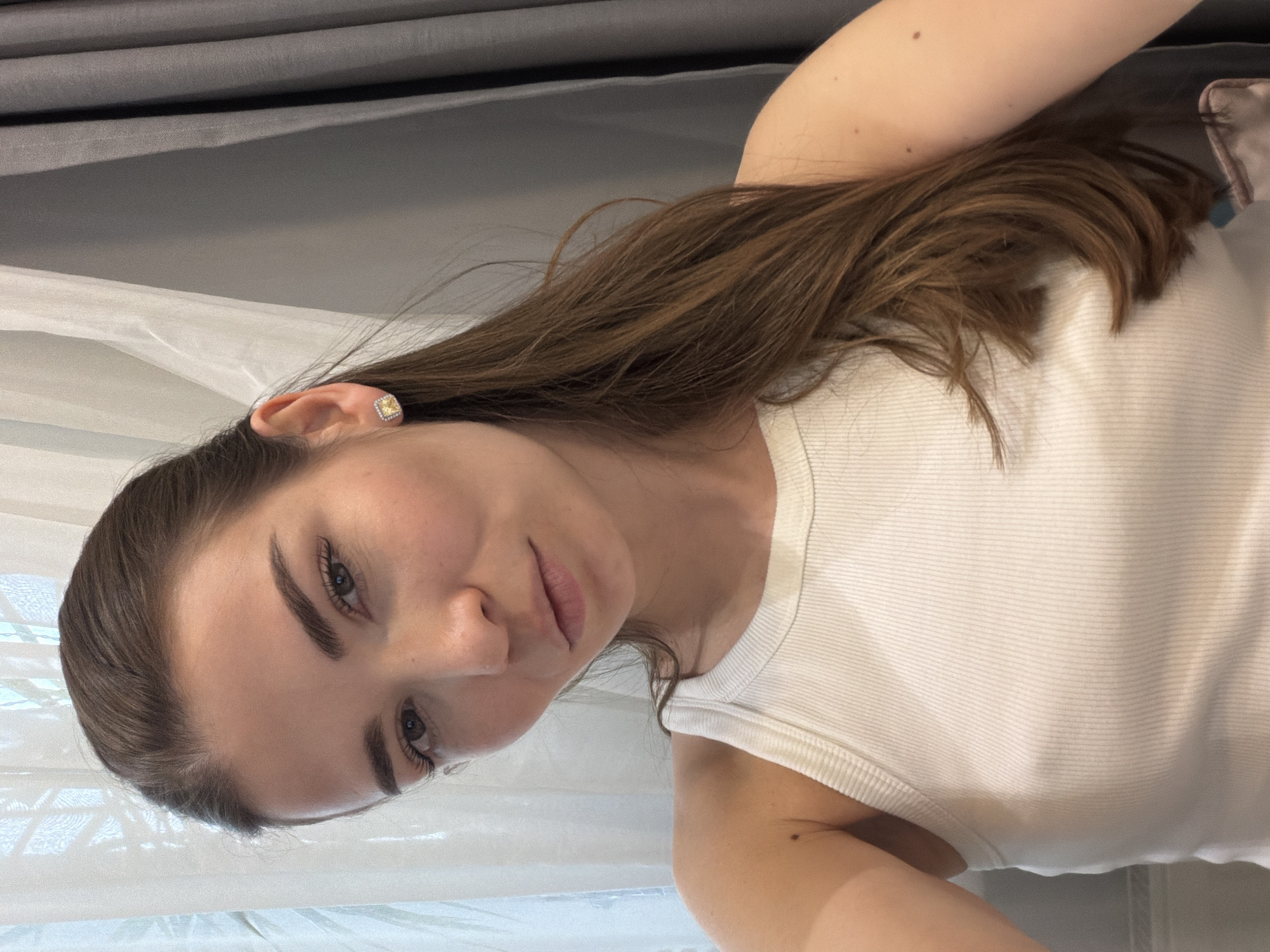 This is a close-up portrait of a young woman, likely taken as a selfie, in Khimki, Russia. The image is oriented sideways, with the woman lying down. She has long, straight dark brown hair, which is fanned out around her. She is wearing a white, ribbed tank top.
Her face is towards the camera, displaying well-defined dark eyebrows, long eyelashes, and light-colored eyes. Her expression is calm and gentle, with a soft gaze directed forward. A square, light yellow stud earring is visible in her left ear. Small moles are present on her visible arm and shoulder.
The background suggests an indoor setting, likely a bedroom, with draped fabrics such as dark grey curtains at the top and sheer white curtains or bedding below. Natural daylight illuminates the scene. There is no visible text.