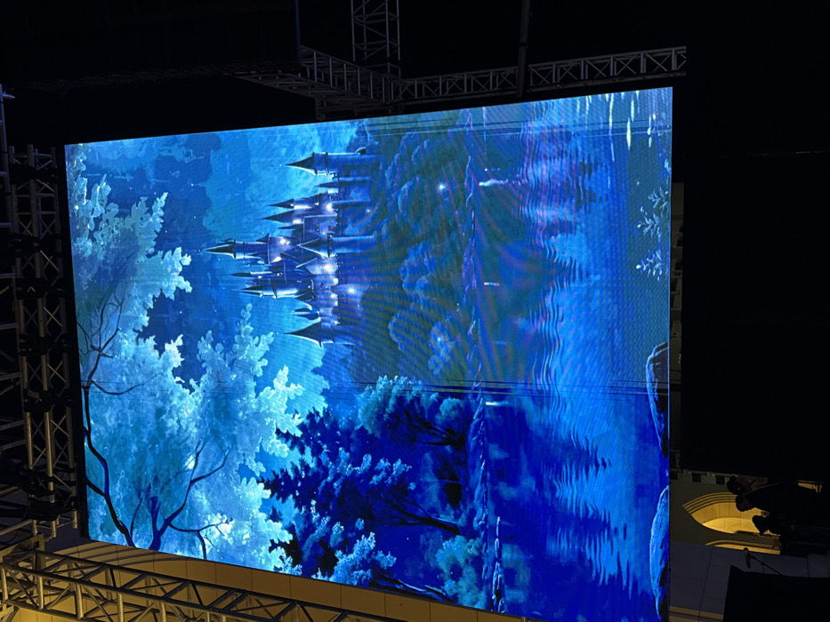 A large digital display shows a fantastical scene dominated by a castle with pointed spires, set amidst a blue-toned forest. The castle glows with light, contrasting with the surrounding dark trees. The right side of the display features a wavy, fluid effect in shades of blue, possibly representing water or a magical distortion. The display is framed by structural supports, hinting at an outdoor event setting.