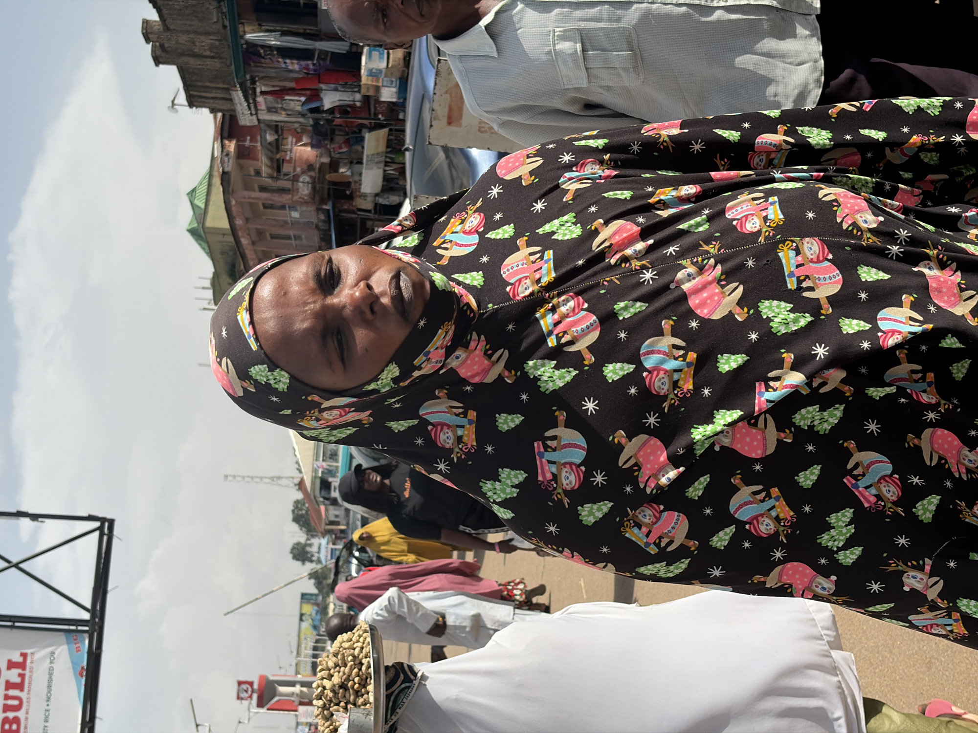 The image captures a street market scene in Jos, Nigeria, with a focus on a woman wearing a black headscarf with a festive sloth pattern. She is looking towards the camera with a neutral expression. Behind her, other market-goers and stalls are visible. One man in the background wears a light blue, button-down shirt. Further back, buildings with awnings and colorful merchandise suggest a busy marketplace. The sky is bright and overcast, indicating it's daytime. In the foreground, a large white object, possibly a table or cloth, occupies the bottom portion of the frame, with a scattering of peanuts in a metal container on its left side. Visible text includes "BULL" and "STY RICE NOURISHED YOU" on a sign, along with "BIG" and other partial text in smaller fonts. The overall impression is a vibrant, everyday scene in a Nigerian market.