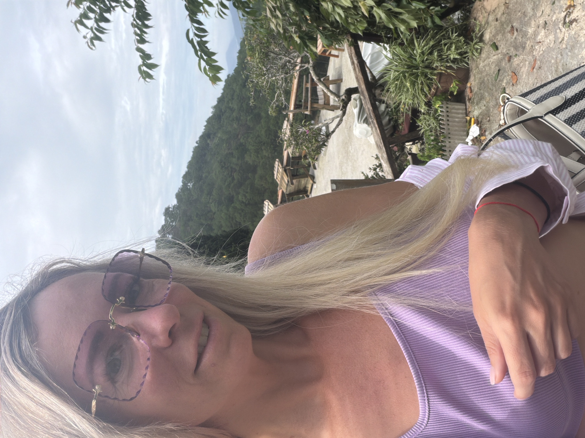 A woman with blonde hair and sunglasses is taking a selfie outdoors. She is wearing a light purple ribbed tank top. Behind her, there is a lush green hillside, a concrete patio with wooden furniture, and some leafy trees. The sky is partly cloudy, suggesting it is daytime. A striped bag is visible on the right side of the frame. The woman appears relaxed and happy.