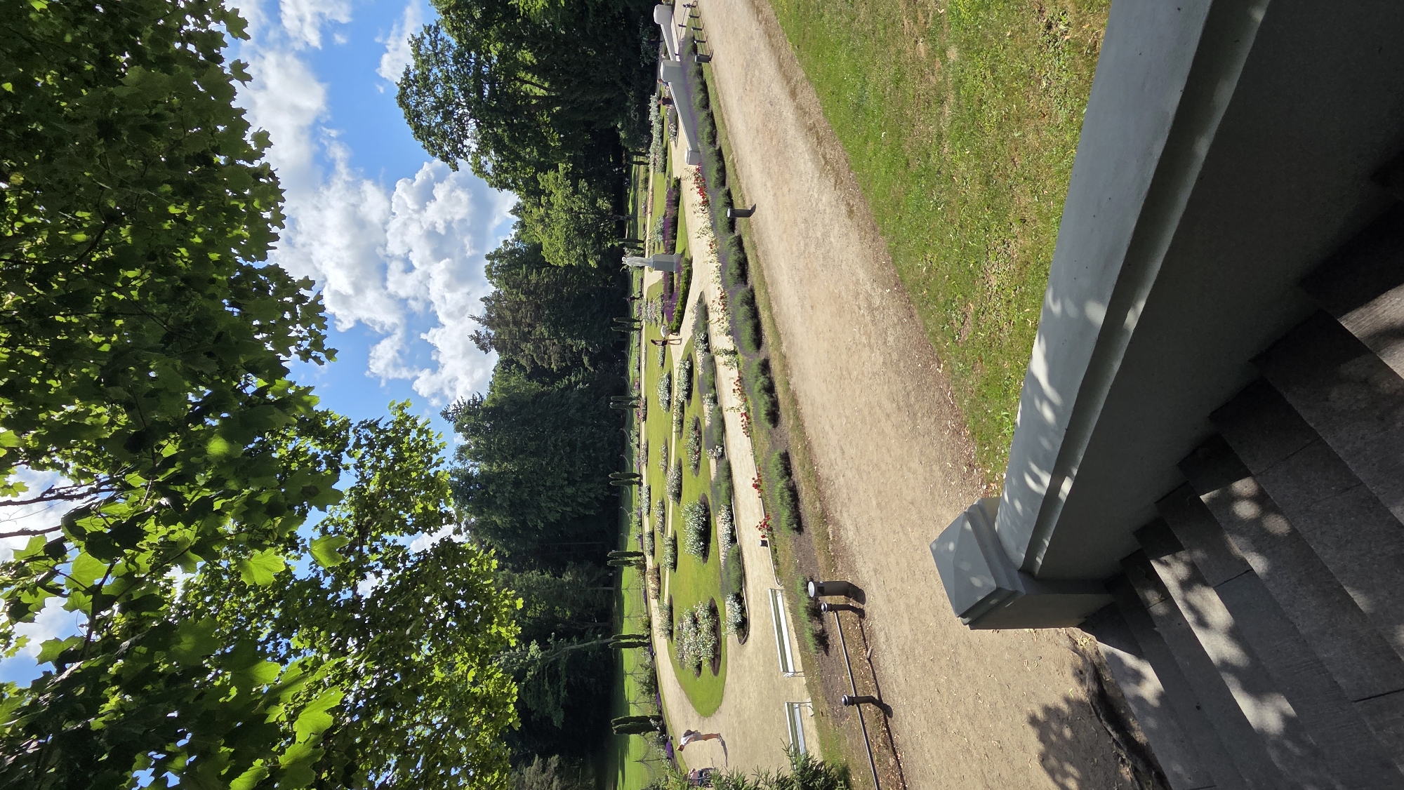 The image depicts a view of a manicured garden with formal flower beds arranged in geometric patterns. The garden is situated in a park with mature trees bordering the flower beds and in the background. Several pathways wind through the garden, and there are white benches along one of the paths. In the foreground, stone stairs and a railing lead down from a vantage point, with dappled sunlight filtering through the leaves of an overhead tree. The sky above is a bright blue with scattered white clouds. A few individuals are visible in the garden, some walking and others standing. This scene is located in the botanical garden of Palanga, Lithuania, specifically the central part of the park with its characteristic landscaping.