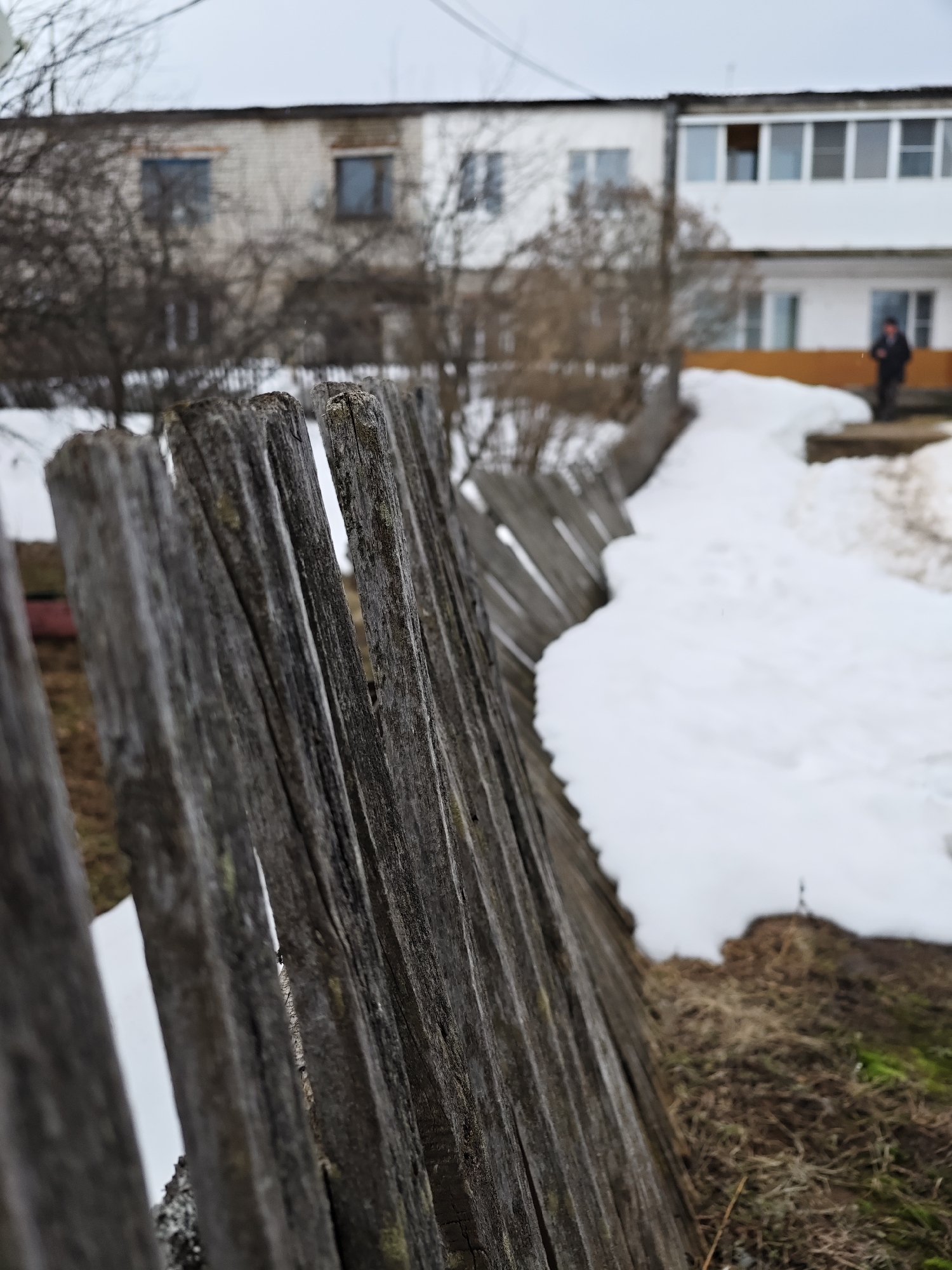 A medium shot shows an aged wooden fence with peeling, gray slats in the foreground, angled towards the right, with gaps in between the slats. A snow-covered area sits beyond the fence, leading up to a two-story building with light-colored walls. A person is vaguely visible standing in front of the building near an orange railing. The sky is overcast, and bare trees are scattered between the fence and the building. Based on the location information, the scene is in Уральское, Russia.