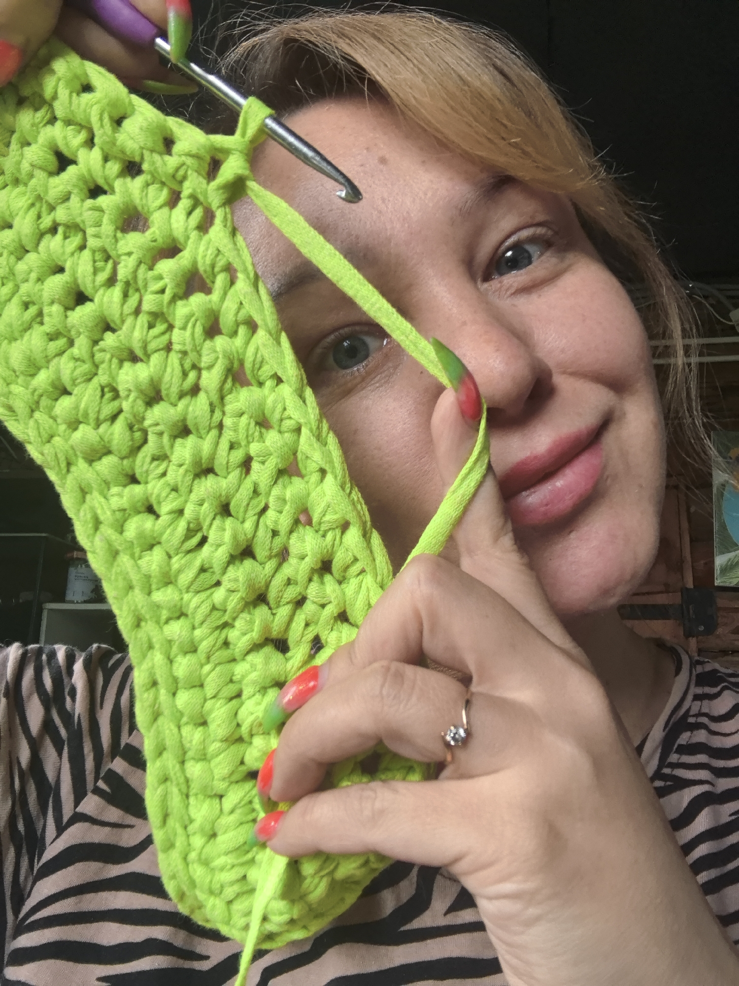 A woman is captured in a close-up, eye-level shot, actively engaged in crocheting a neon green fabric item. Her blonde hair frames her face, and her blue eyes are looking directly at the viewer with a friendly smile. She is holding a silver crochet hook with a purple handle, which is inserted into the bright green yarn, indicating an ongoing stitch. Her fingers, adorned with vibrant red and green gradient nail polish, are deftly manipulating the yarn and the hook. On the ring finger of her right hand, a delicate gold ring with a single clear stone is visible.
The crocheted item is made from thick, brightly colored yarn, forming a dense, textured pattern. Part of a zebra-print garment is visible on the woman's torso, suggesting she is wearing a striped top or is seated near a patterned fabric. The background is softly blurred but hints at an indoor setting, possibly a home or craft area, with dark and light elements, including what appears to be shelves or furniture. The lighting suggests it is daytime. The woman's expression conveys contentment and focus on her craft. The activity takes place in Noviy, Russia. There is no visible text in the image.