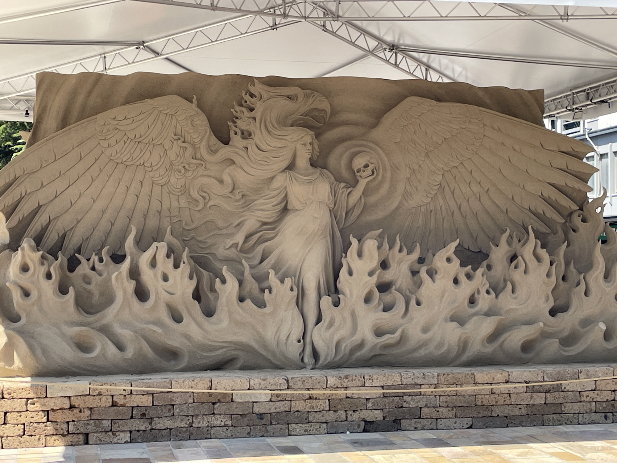 The image shows a large sand sculpture in Jesolo, Italy, depicting a woman with large feathered wings and an eagle's head emerging from flames. The woman holds a skull in her right hand. The sculpture is set against a backdrop of a tent structure and brickwork.