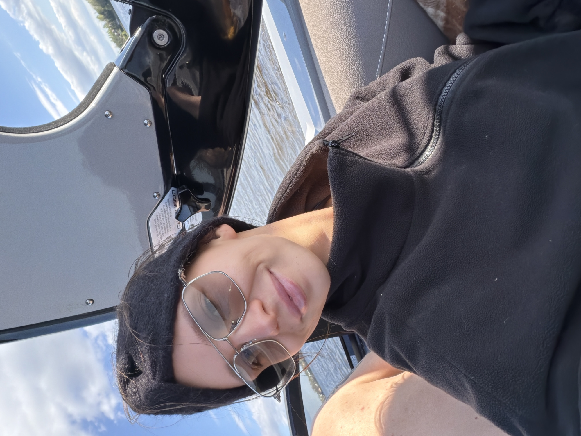 A woman wearing a black hooded sweatshirt and silver-rimmed glasses is taking a selfie on a boat. She is wearing a black headband. Part of the boat's console is visible to the left. The sky and water are visible in the background. The specific location is not discernible from the image.