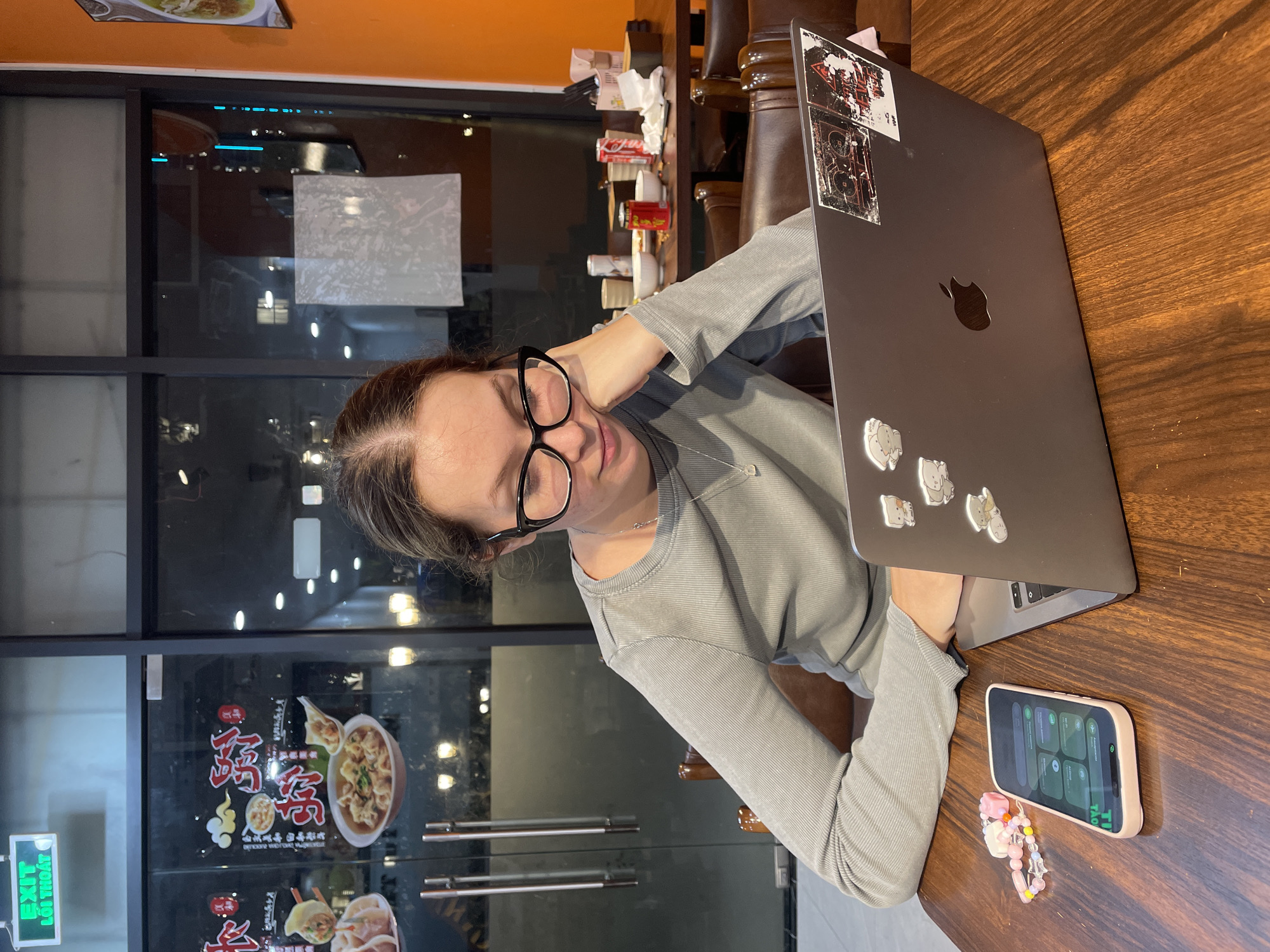 A woman with dark hair, wearing glasses and a gray long-sleeved shirt, is seated at a wooden table with a laptop and a smartphone. She is resting her head on her hand, appearing to be tired or thoughtful, as she looks towards the camera. The laptop, adorned with several stickers, is open on the table. A pink-cased smartphone, displaying a grid of app icons, lies next to a charm bracelet. The background suggests an indoor setting, possibly a restaurant or cafe, with large windows reflecting external lights and street scenes. Signs in Vietnamese, including "EXIT" with "LỐI THOÁT" below it, are visible on the glass. The overall lighting indicates it is likely evening or night. The presence of food imagery on the window also suggests a dining establishment.