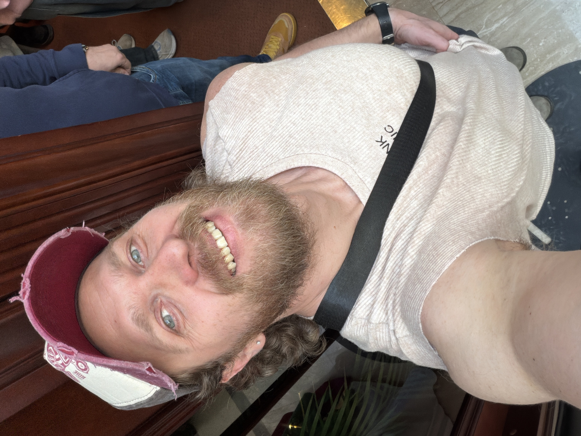 A selfie of a bearded man with blue eyes and a distressed baseball cap smiling at the camera. He is wearing a light-colored ribbed shirt and a black strap crosses his chest. The photo also captures glimpses of other people, including one wearing jeans and a blue sweater and another with yellow shoes. The backdrop appears to be an interior setting, possibly a restaurant, with a shiny floor and brown furniture. There is no information to identify the location as Saint Petersburg, Russia.