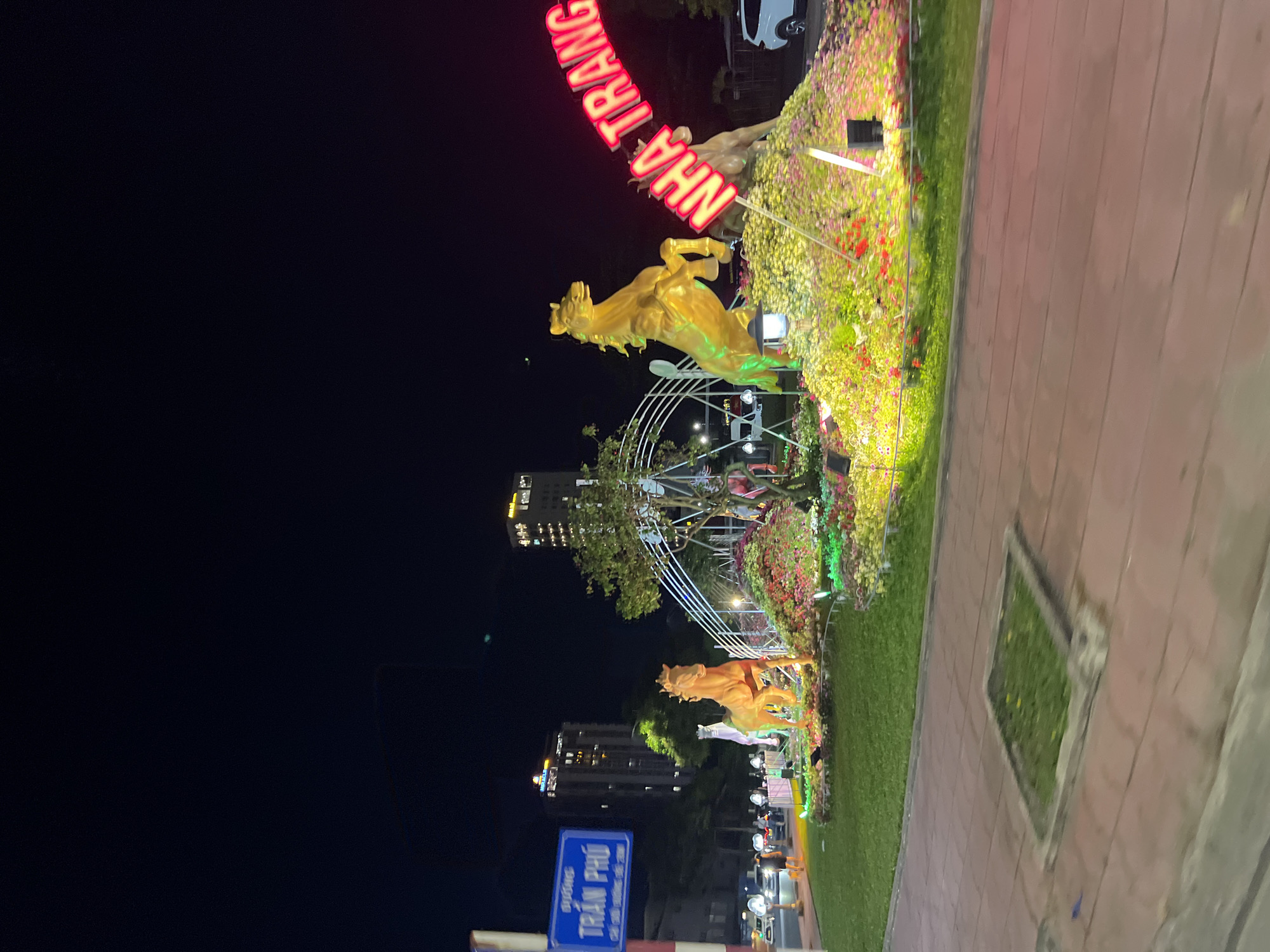 At night, the Nha Trang street art installation displays golden horses, flowers, and neon signs spelling out "NHA TRANG". Street sign reads "TRÁN PHÚ" . Buildings and street lights are visible in the background. A paved sidewalk borders a grassy area.