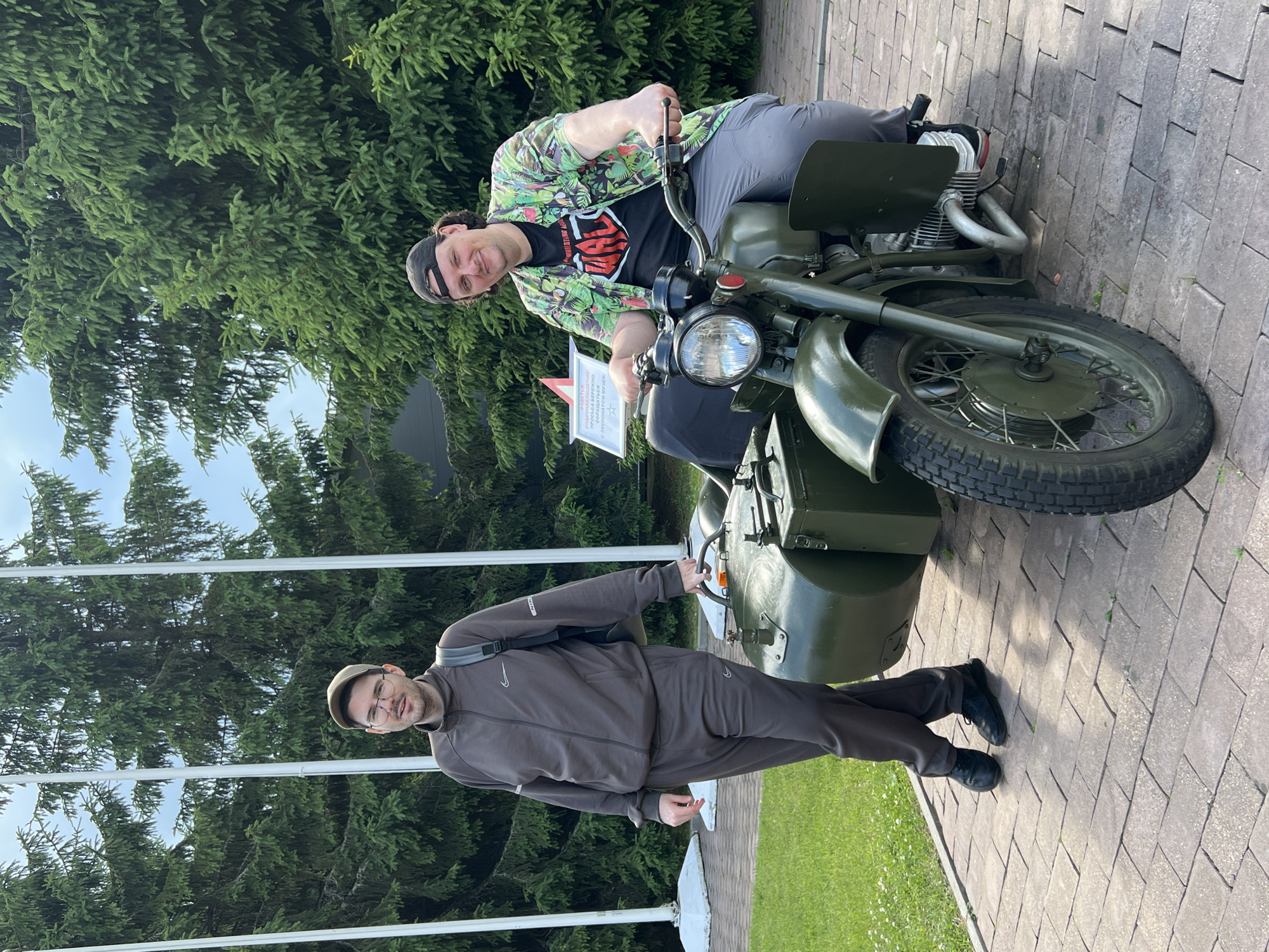 Two men are posing with a vintage green military motorcycle with a sidecar. The man in the foreground is wearing a brown tracksuit, a cap, and glasses, and is standing next to the motorcycle. He is looking at the camera with a neutral expression. The man in the background is sitting on the motorcycle and wearing a black t-shirt with a red and white logo, a brightly patterned shirt, and a black baseball cap. He is also looking at the camera with a slight smile. The motorcycle appears to be an older model, possibly from the Soviet era, given the location context of Kubinka, Russia, which is known for its tank museum. The setting is outdoors, with a paved walkway and lush green trees in the background. The sky is visible and appears bright, suggesting it is daytime. There are white poles with flags or banners visible behind the man in the foreground. A small sign with Cyrillic text is visible next to the motorcycle, but the text is too small to be legible. The overall mood appears casual and friendly.