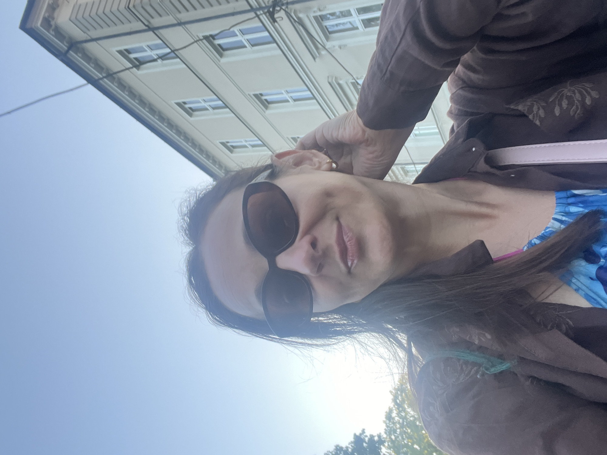 A medium shot captures a woman with dark hair wearing sunglasses and a brown jacket. She is touching her ear with her right hand, which is positioned behind her head. A portion of a light-colored building with multiple windows is visible in the background, along with a clear blue sky. The woman is looking towards the camera.