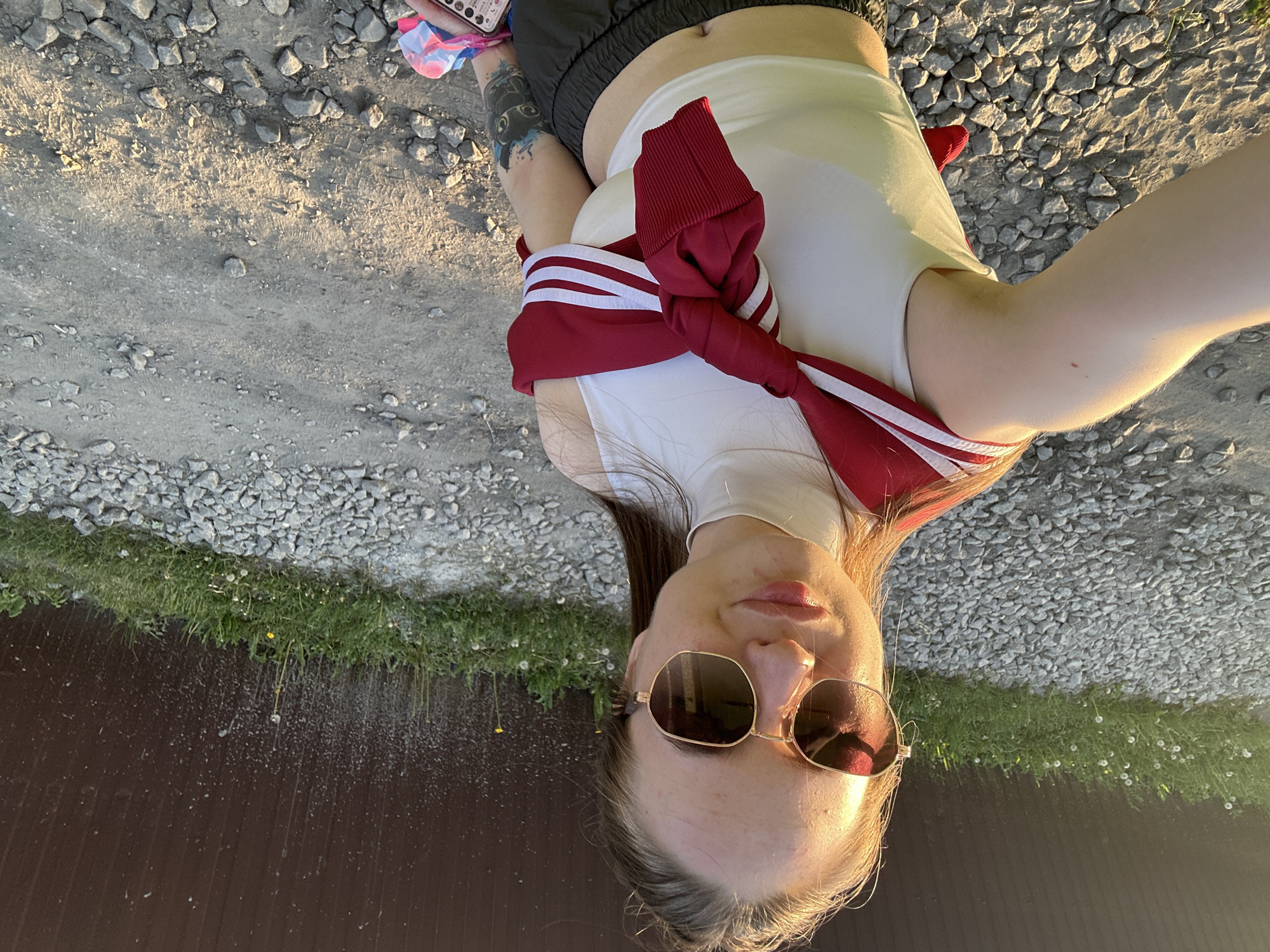 The image depicts a young adult female taking a selfie outdoors in Unknown, Unknown. She is positioned centrally, holding the camera slightly above eye level, capturing herself from the chest up and some of the immediate surroundings. The ground is a mix of light grey gravel and packed dirt. In the background, there is a dark brown, corrugated metal fence or wall, with a strip of green grass and scattered white dandelions growing at its base.
The female subject has fair skin and long, straight, dark brown hair that appears pulled back. She is wearing large, hexagonal-framed sunglasses with gold metallic rims and dark, reflective lenses. Her lips are slightly parted in a subtle smile. She wears a white, sleeveless, mock-neck crop top. Around her shoulders, a maroon or dark red long-sleeved sweatshirt with two white horizontal stripes on each sleeve is tied, forming a knot at her chest. Her bare midriff and belly button are visible. A prominent tattoo featuring a dark, stylized animal head (possibly a cat or fox) with blue and teal accents is visible on her left forearm/upper arm. On her right bicep, a small reddish mole is visible. Her left hand, partially visible at the bottom right, holds a white smartphone with multiple camera lenses and a colorful, patterned fabric item, possibly a scrunchie, in pink, blue, and white. The scene is illuminated by warm, golden sunlight, indicating either late afternoon or early evening, with strong highlights on the subject's right side (viewer's left).