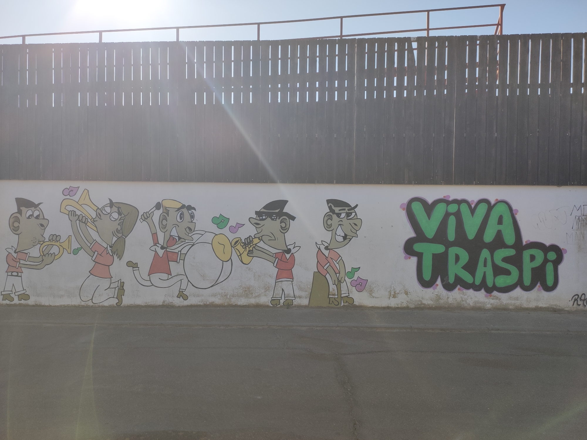 The image depicts a section of a white wall covered in graffiti, located in Traspinedo, Spain. On the left side of the wall, a cartoon band is painted, featuring five characters playing various musical instruments. The characters are depicted in a simple, playful style. To the right of the band, the words "VIVA TRASPI" are spray-painted in large green lettering with a dark outline. The wall is at ground level, and the foreground shows a grey asphalt surface. Above the wall, a dark wooden fence with vertical slats is visible, and a clear blue sky is in the background. Sunlight casts lens flares across the image.