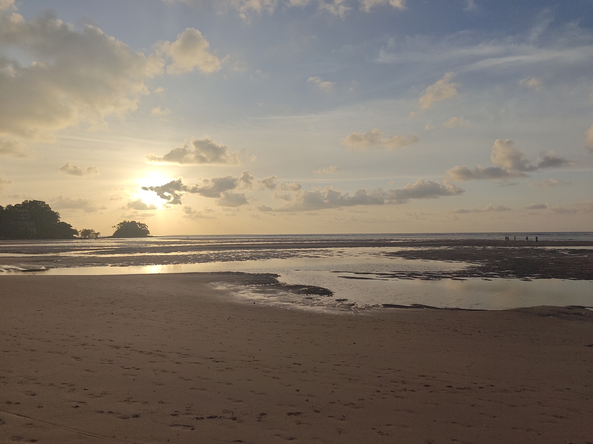 The image depicts a serene beach scene at low tide in Sakhu, Thailand, during either sunrise or sunset. The sun, a bright orb, is partially obscured by clouds and casts a warm, golden light across the sky and the water. The sky is a gradient of soft blues and yellows, dotted with scattered white and gray clouds.
In the foreground, a sandy beach stretches across the frame, marked by numerous footprints and tire tracks. The sand appears damp in places, suggesting the tide has recently receded. Beyond the beach, shallow pools of water reflect the sky and the distant landscape.
Further out, the sea stretches to the horizon, a calm expanse of water. A small island, covered in lush green trees, is visible in the distance to the left. On the right side of the image, a group of five people are seen walking along the water's edge, their figures silhouetted against the bright sky. The overall atmosphere is peaceful and tranquil, evoking the natural beauty of the coastal environment.