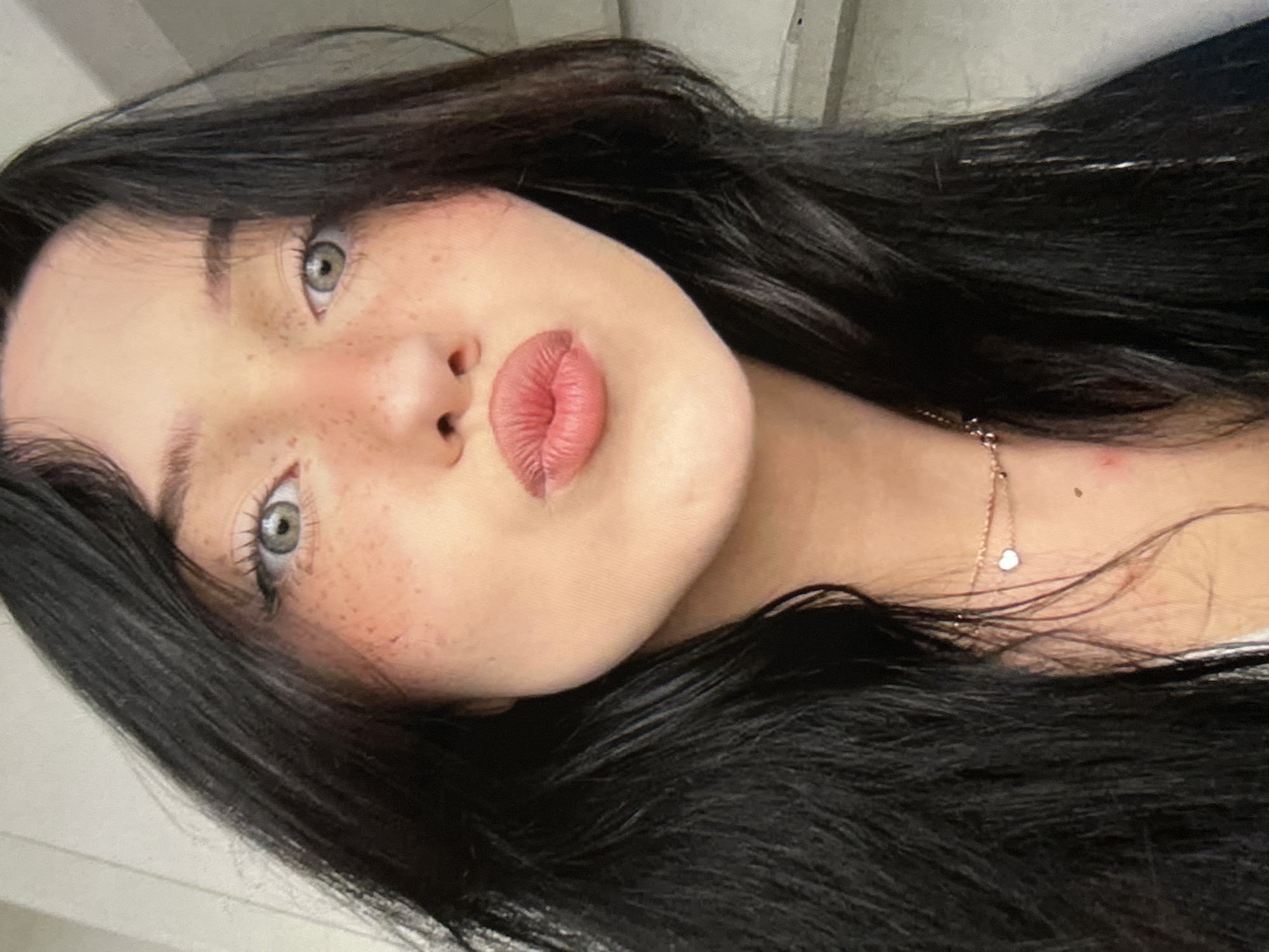 A close-up, head-and-shoulders shot shows a young woman with long, dark hair and light-colored eyes. She has freckles scattered across her cheeks and nose, and her lips are pursed in a kissing gesture. She is wearing a delicate necklace with a small heart pendant. The background is indistinct and appears to be an indoor setting with a neutral-colored wall. There is no indication of the city of Cupra Marittima, Italy, or any specific environmental details that would place the image in that location.