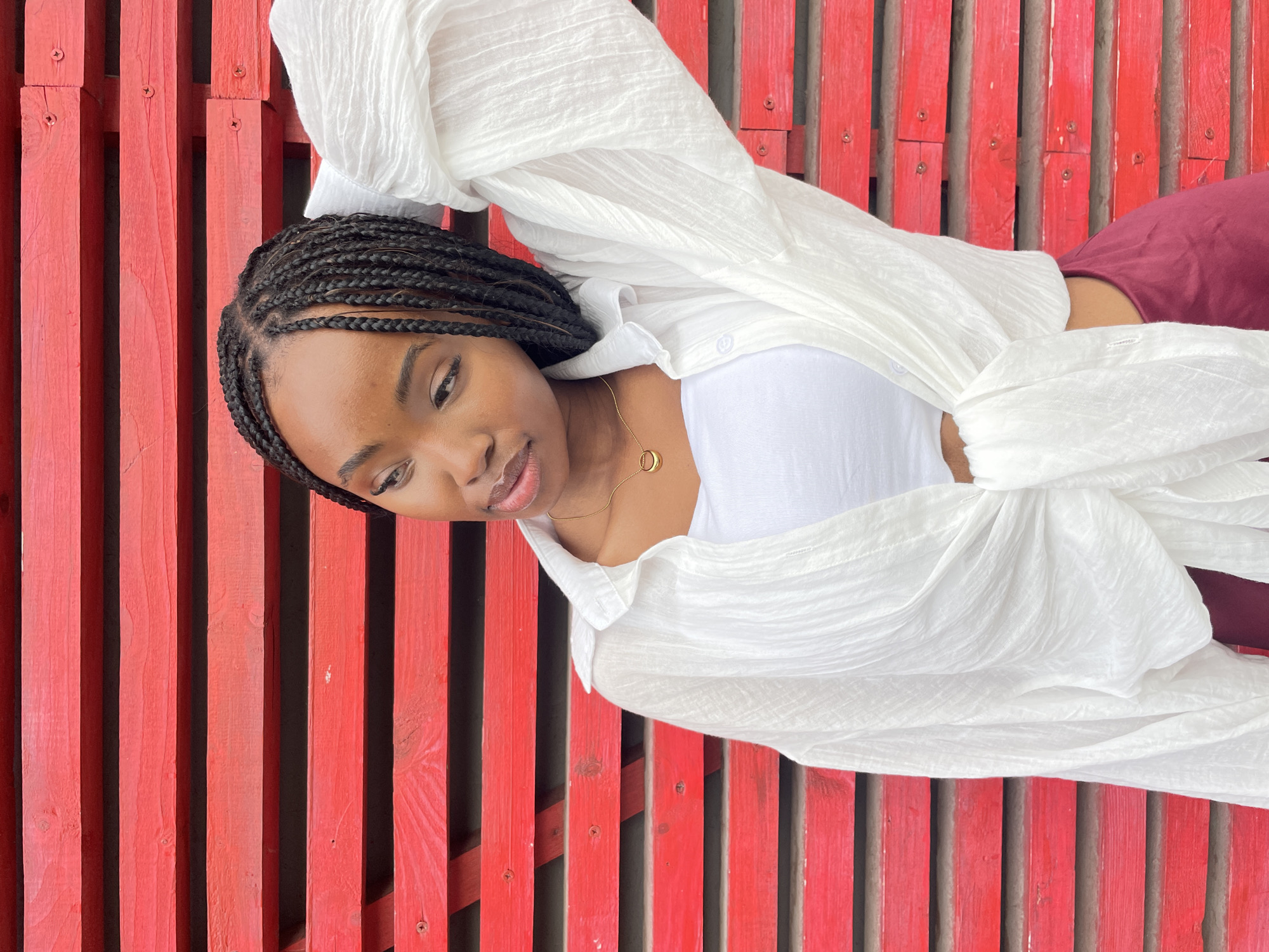 A young Black woman with braided hair stands in front of a bright red slatted wall. She is wearing a white crop top and an oversized white button-down shirt, the front of which is tied at her waist. She has a gold pendant necklace. Her right arm is raised, with her hand behind her head. The background appears to be an exterior wall made of horizontal red wooden planks. There are visible screws fastening the planks. The lighting suggests it is daytime. The image was taken in Kazan, Russia.