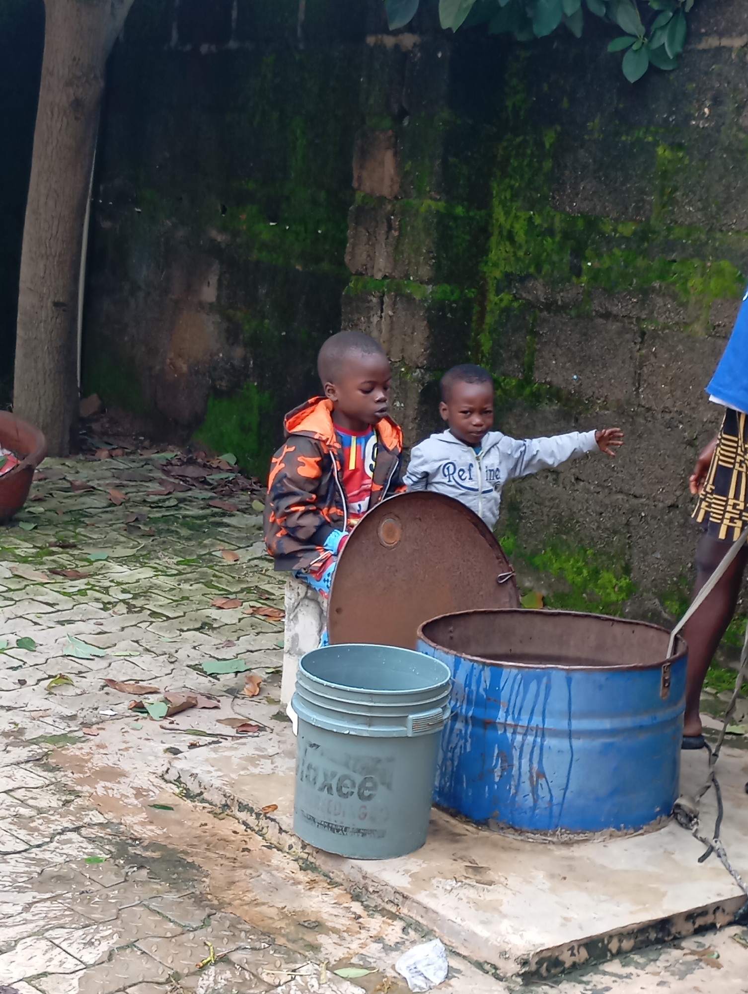 Two young boys, likely brothers, are standing next to a large, blue metal barrel filled with water. The barrel is situated on a concrete platform outdoors. A gray plastic bucket with the word "axee" visible on its side is placed next to the barrel. A rusted metal lid for the barrel is lying on the ground nearby.
The older boy, on the left, is wearing a camouflage-patterned jacket over a colorful shirt and blue patterned shorts. He is looking down at something in front of him. The younger boy, on the right, is wearing a light gray hooded sweatshirt with the word "Refine" visible on it. He is pointing his right arm outwards, as if indicating something. The children appear to be engaged in some sort of activity or game involving the barrels and buckets.
In the background, a moss-covered brick wall suggests an outdoor setting that may experience significant rainfall. The ground is paved with interlocking stones and is wet, with fallen leaves scattered across it, indicating it might have recently rained or is in a humid environment. A portion of a tree trunk is visible on the left side of the frame. A person's legs, wearing a black and yellow patterned skirt and dark shoes, are visible on the far right, suggesting an adult is present, possibly supervising the children.
The overall impression is of a casual, everyday scene in a residential or communal outdoor space in Jos, Nigeria. The lighting suggests it is daytime, and the damp ground hints at recent precipitation or high humidity common in the region. There are no strong emotional cues from the children, but their engagement with the objects implies curiosity and playfulness.