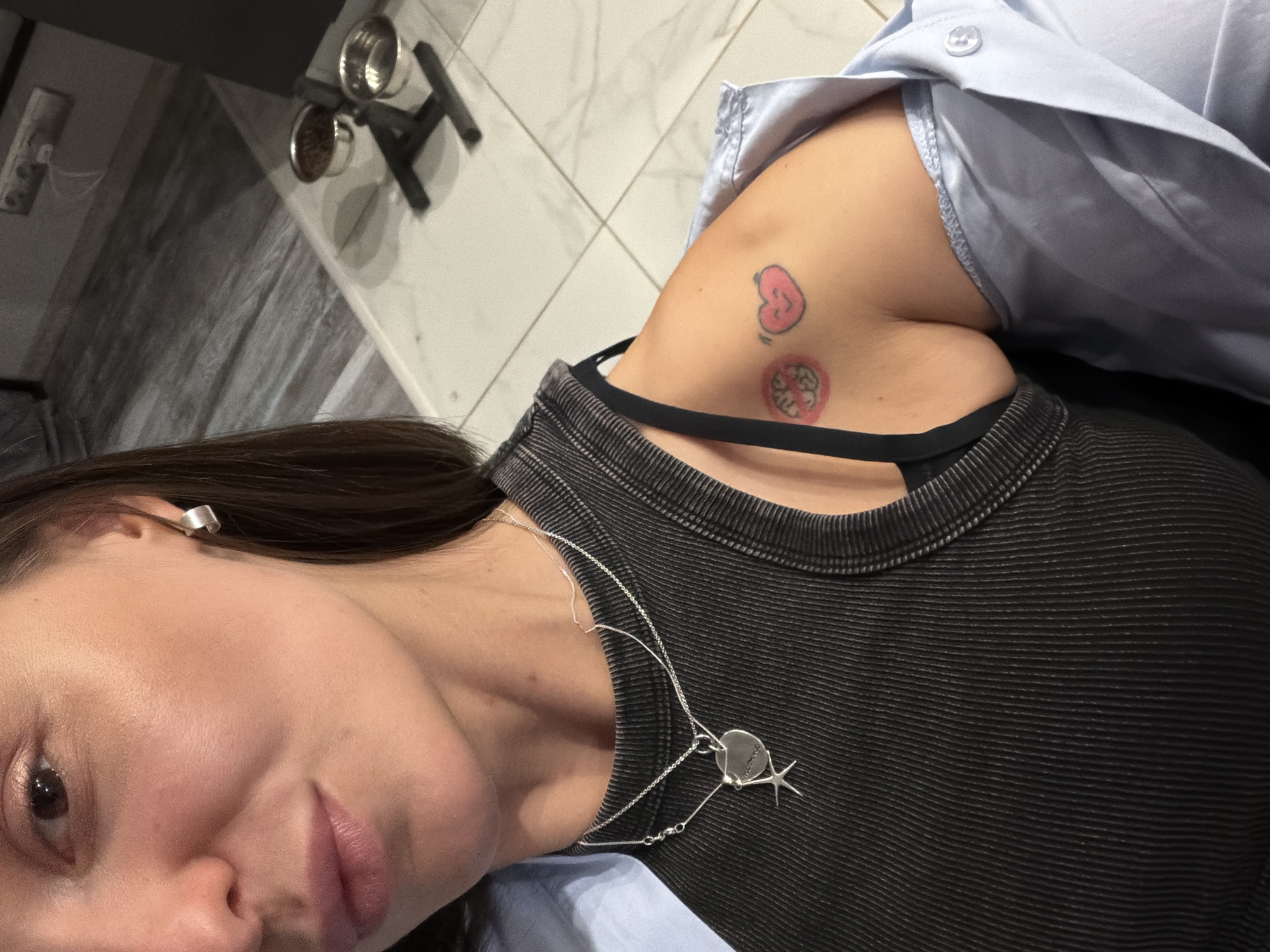 A selfie taken from a high angle depicts a fair-skinned individual with dark hair, wearing a black top with horizontal ribbing and a light blue collared shirt. A silver necklace with a pendant is visible around their neck. On their right shoulder, two tattoos are present: one is a simple heart outline filled in with pink, and the other is a pink circular design. An ear cuff is worn on the left ear. The background includes a section of gray patterned wall and a small metal structure with dishes. The location is not discernible in this image, therefore, the assertion that this is Moscow, Russia cannot be confirmed.