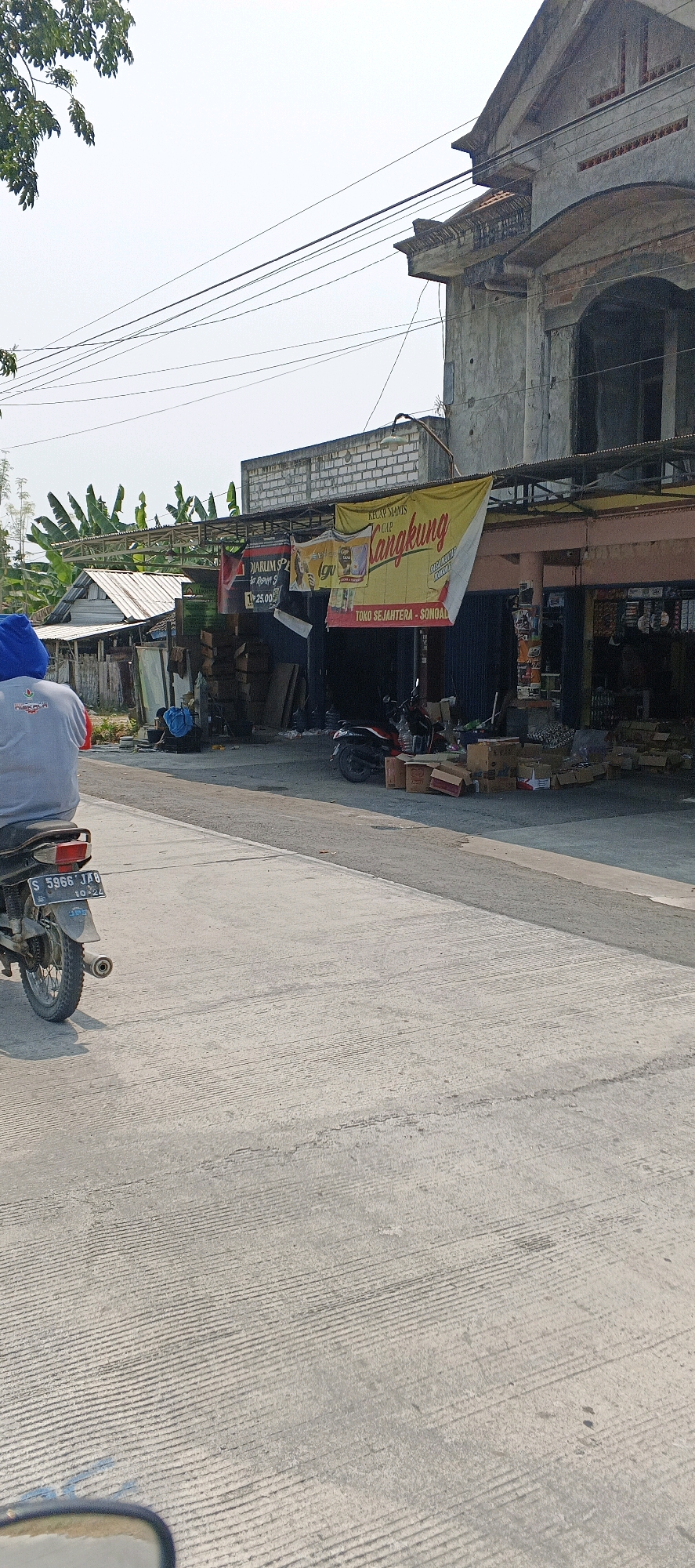 The image captures a street scene in Guci, Indonesia, likely a commercial area. In the foreground, a person on a motorcycle, viewed from behind, is on the right side of the road. The motorcycle has a license plate that reads "S 5966 JAB". The road is paved with concrete, showing visible lines from its construction. To the left, a person wearing a blue head covering and a grey shirt is seated on the motorcycle.
The background features a row of shops and buildings. One prominent structure is a building with a partially constructed or dilapidated facade, made of concrete and brick, with exposed rebar and an arched window. In front of the shops, a yellow banner with Indonesian text reads "KECAP MANIS LANGKUNG" and below that, "TOKO SEJAHTERA - SONDA". Several other banners and signs are visible, including one with red lettering and numbers possibly indicating prices. Boxes and merchandise are stacked outside the shops, and two motorcycles are parked near the storefronts. Behind the shops, banana trees are visible, suggesting a tropical or subtropical environment. The sky is bright and overcast.