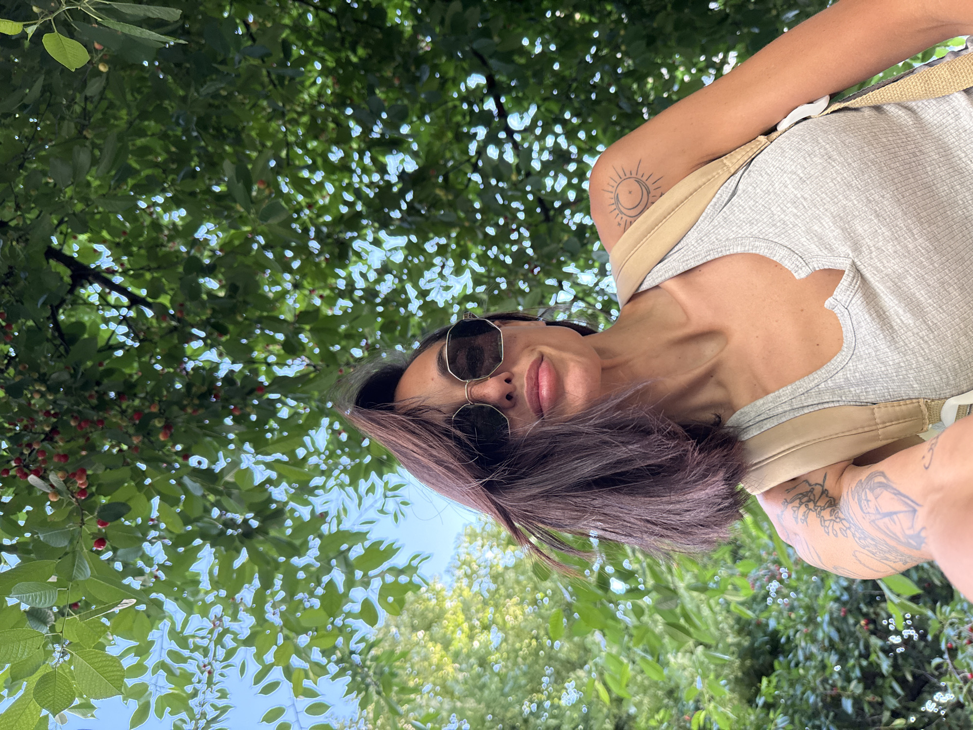 A low-angle, waist-up selfie shows a woman with dark brown hair and sunglasses posing outdoors. She is wearing a grey ribbed tank top and a tan backpack. A tattoo of a crescent moon with sun rays is visible on her right shoulder. The background is filled with the green leaves and red cherries of a tree, with dappled sunlight filtering through. The scene appears to be in a park in Pleven, Bulgaria.