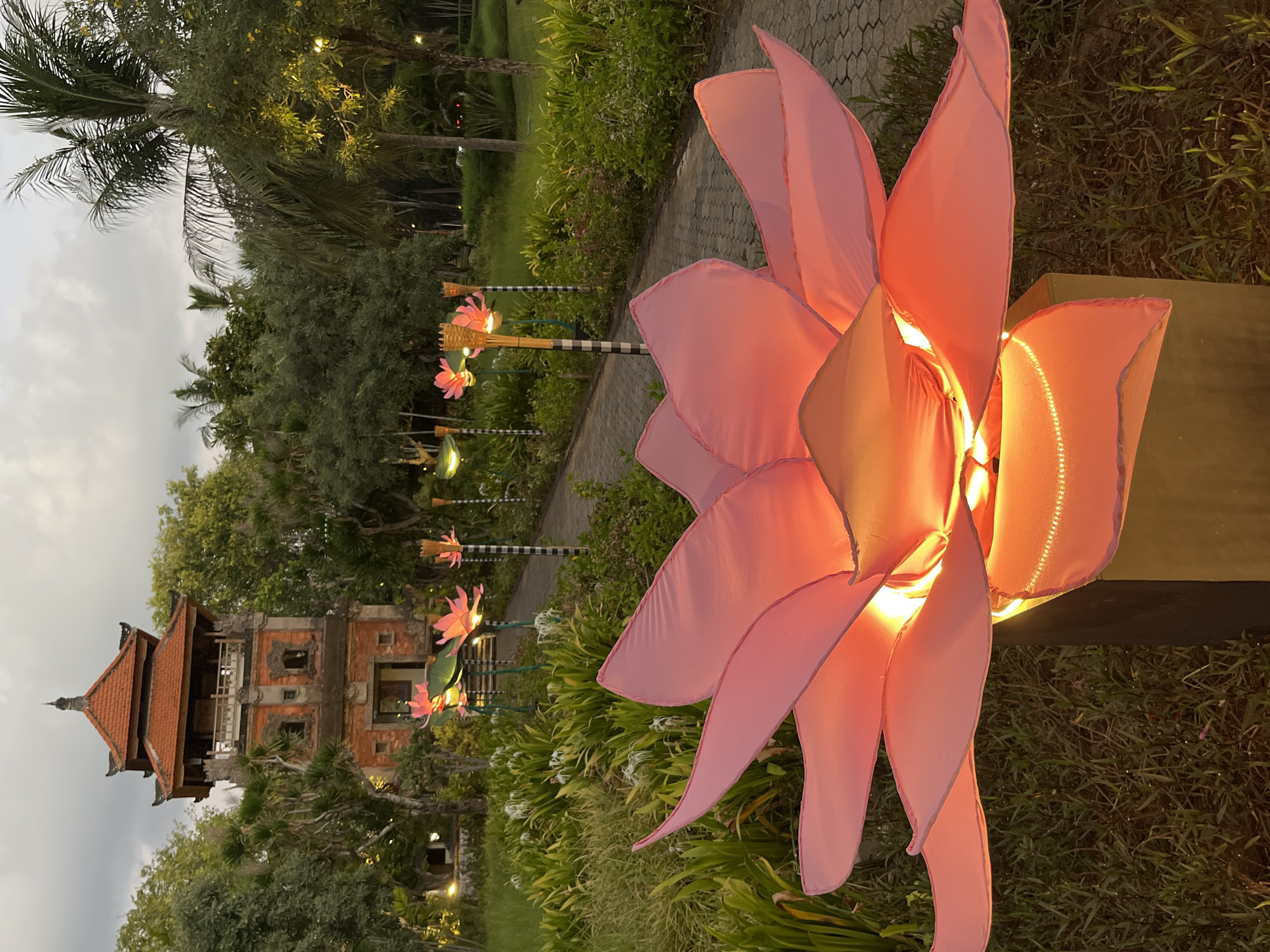 In Kuta Selatan, Indonesia, a nighttime scene unfolds in a park adorned with illuminated lotus flower decorations. In the foreground, a large, pink, fabric lotus flower sculpture, lit from within with a warm orange glow, dominates the frame. Its petals are wide and gently curved, some detailed with subtle LED string lights. Behind this prominent flower, a paved walkway curves through the park, lined with more, smaller illuminated lotus flowers on tall poles. These flowers are also pink and lit from within. The walkway leads towards a traditional Balinese tower, made of brick and red tile, nestled amongst lush green trees and palm trees. The sky above is overcast and grey, suggesting dusk or a cloudy evening. The overall atmosphere is serene and decorative, with the illuminated flowers creating a soft, inviting ambiance.