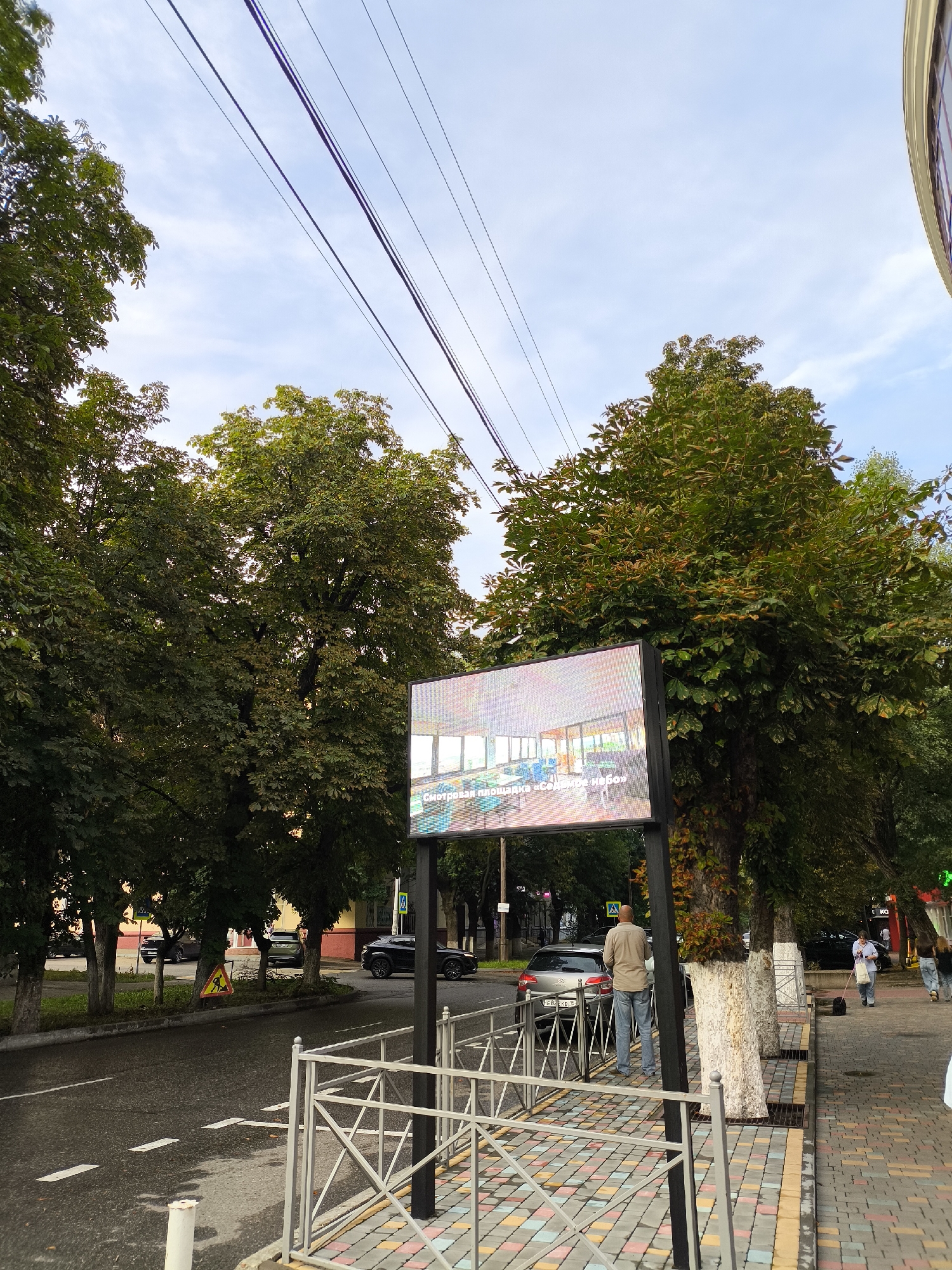 The image depicts a street scene in Yessentuki, Russia. In the foreground, a colorful tiled sidewalk is visible, bordered by a white metal fence. A large digital billboard displays an interior scene of a well-lit room with modern furniture. A man stands near the billboard, facing away from the camera. Behind him, a street runs alongside the sidewalk, with cars parked and driving on it. Lined with trees, the street leads to buildings in the background. The sky above is partly cloudy, with visible power lines stretching across the frame. The overall impression is of a commercial or public area in a Russian city.