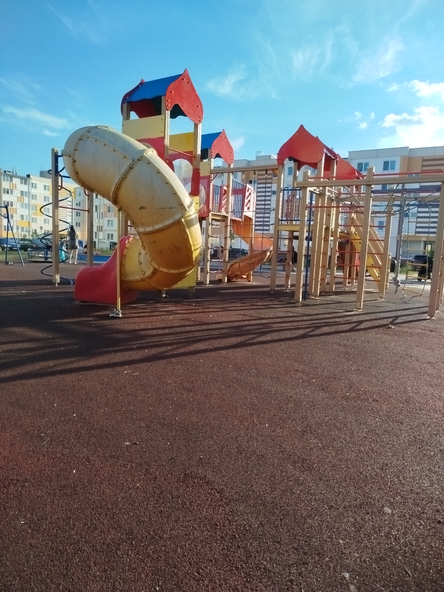 The image depicts a playground with a large, multi-colored slide structure. The slide is predominantly yellow and red, with a red lower section. To the left of the slide, there are apartment buildings visible, suggesting an urban or suburban setting. The ground of the playground is covered with a dark brown, rubberized surface. The sky is blue with scattered white clouds. In the background, there are several people on the playground, including a woman with a child, and some parked cars. The scene is illuminated by natural sunlight, casting shadows on the playground surface.