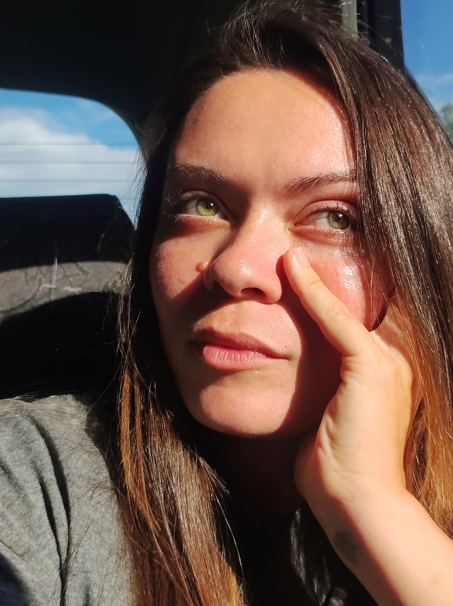 A close-up, eye-level, over-the-shoulder selfie shows a young woman with long, brown hair. She is sitting in the passenger seat of a vehicle, with the window on her left revealing a blue sky with white clouds and the interior of the car on her right. Sunlight illuminates her face from the left, casting shadows and highlighting her green eyes. Her right hand is raised to her face, with her index finger touching her cheekbone. She is wearing a grey t-shirt. A faint tattoo is visible on her wrist. The setting appears to be Chaikovsky, Russia, based on the provided context.