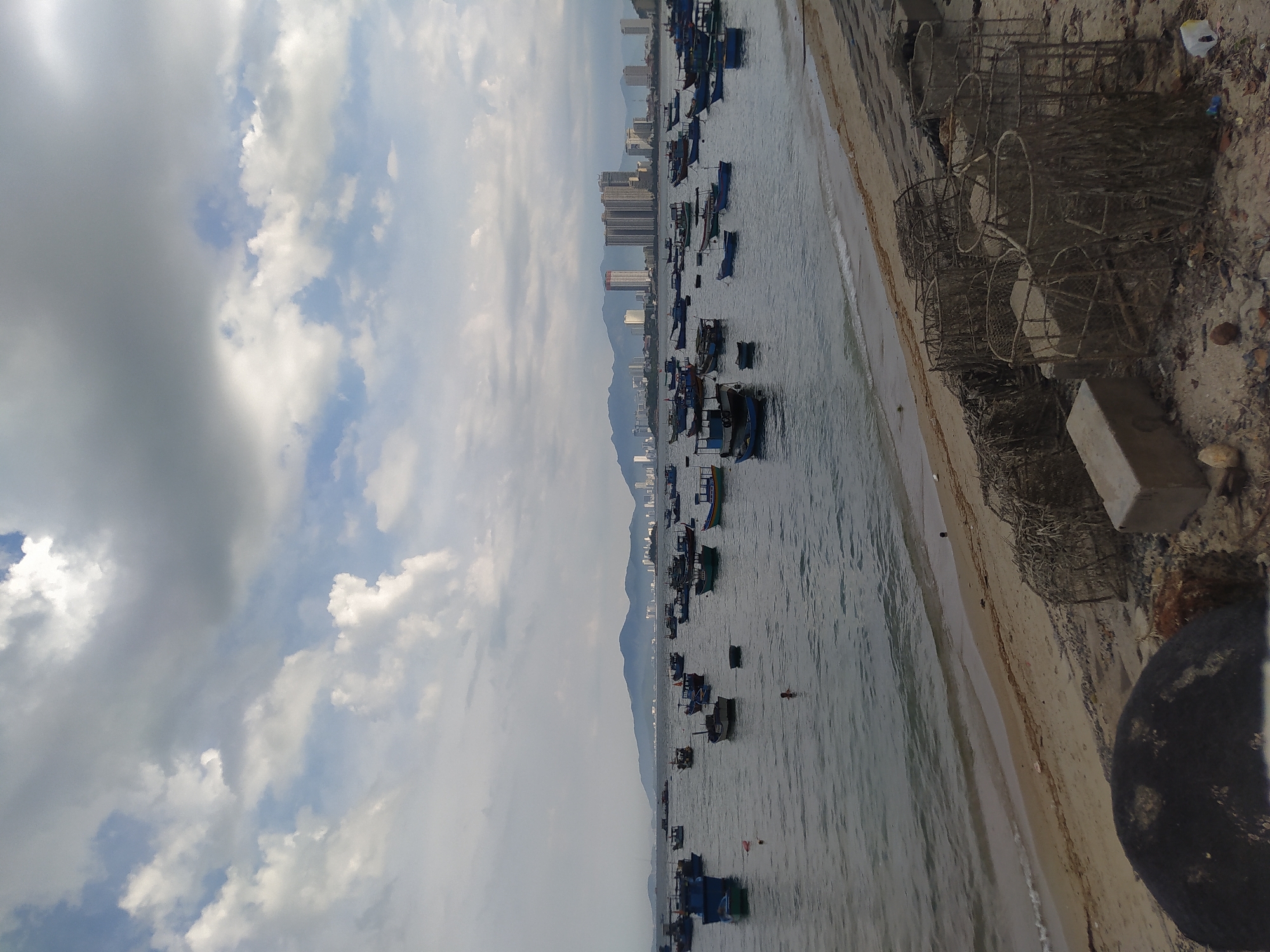 The image shows a body of water with numerous small boats anchored or moored. A sandy beach is visible to the right of the water, with fishing traps and debris scattered along the shore. In the distance, a city skyline with tall buildings rises against a backdrop of mountains. The sky is partly cloudy, with patches of blue peeking through. The scene appears to be a coastal area, possibly a bay or harbor, with the city suggesting a populated region. The time of day is unclear, but the lighting suggests it could be morning or late afternoon. There are no people visibly engaged in activities, though one figure can be seen in the water, possibly swimming. The overall mood is serene and quiet, with a sense of natural beauty juxtaposed with urban development. The image is oriented vertically, suggesting it was captured from an elevated perspective looking down at the scene.