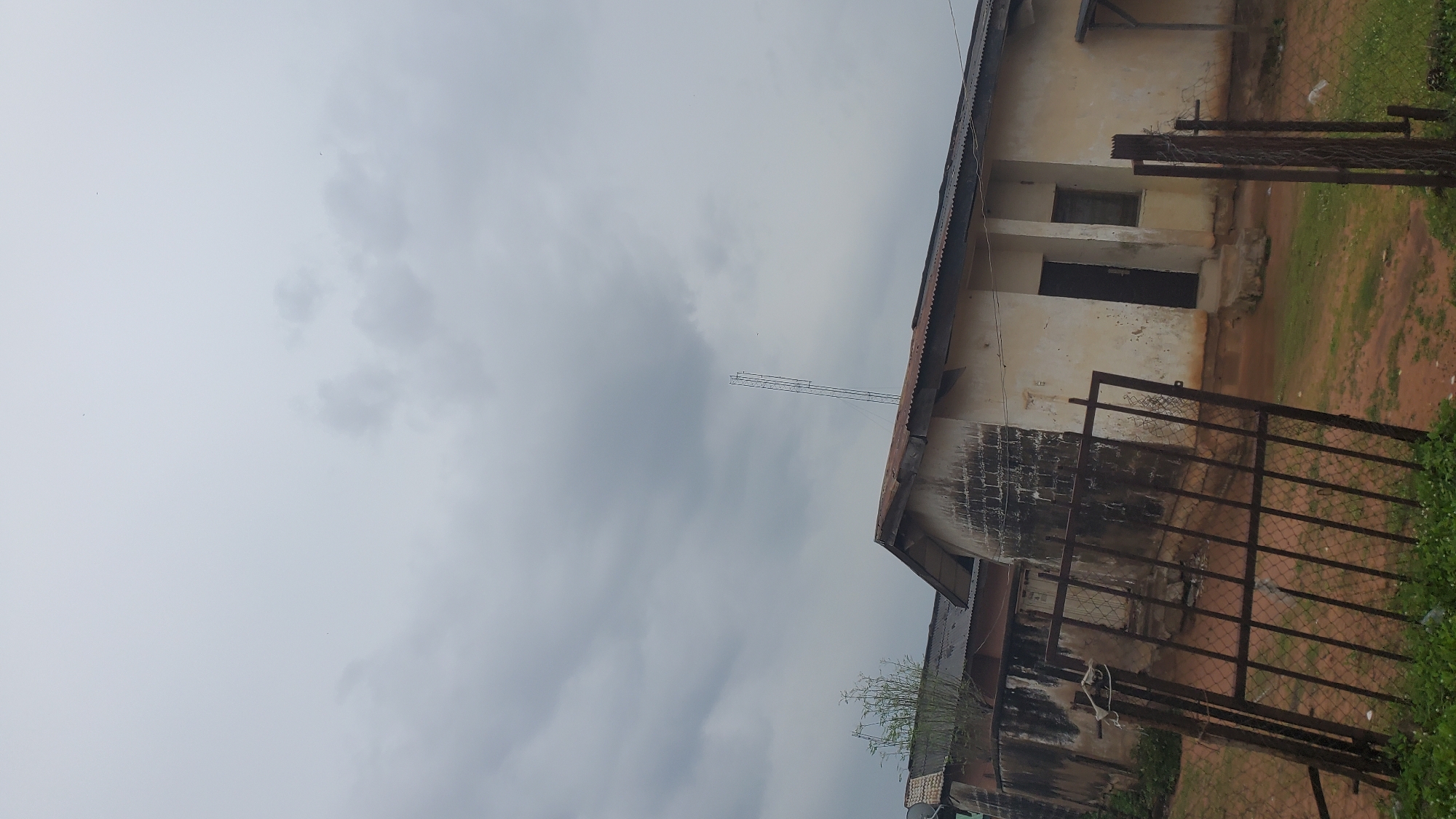 The image shows a cloudy sky over a modest building in Jalingo, Nigeria. The building appears to be an older structure with a weathered, off-white facade and a corrugated metal roof. It has at least one door and a small window visible. In the foreground, a rusty metal gate with a diamond-shaped mesh pattern stands in front of the building. The ground is covered in dirt and sparse green vegetation. A tall metal tower, possibly a communication mast, is visible in the distance behind the building. There are no people visible in the scene.