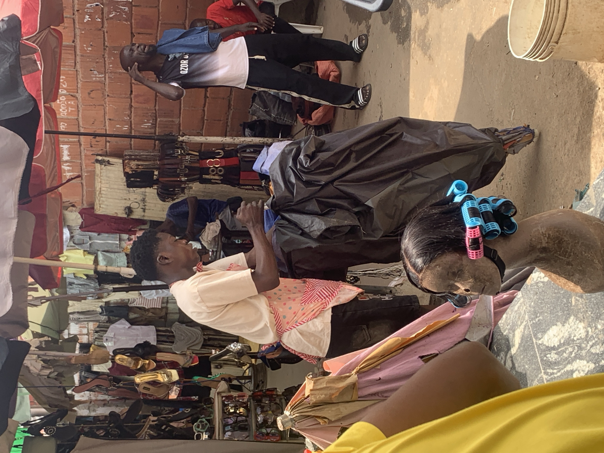 The image captures a scene in a bustling outdoor market in Abuja, Nigeria. In the foreground, a mannequin head with curlers in its hair is visible, suggesting a hair salon or beauty product vendor. To the left, a person wearing a yellow shirt is partially visible. In the center, a man wearing a light-colored t-shirt with a patterned apron over it is attending to a large black plastic-wrapped bundle. The background reveals a vibrant market setting with stalls displaying clothing, shoes, and accessories, as well as a brick wall. Another man, wearing a dark t-shirt and dark pants with a stripe down the side, is standing to the right, talking on a phone and holding a denim jacket over his shoulder. A man in a red shirt is seated on a chair in the background. The ground is dirt and sand, typical of an outdoor market. The overall atmosphere is lively and suggestive of commerce and daily life in a Nigerian city.