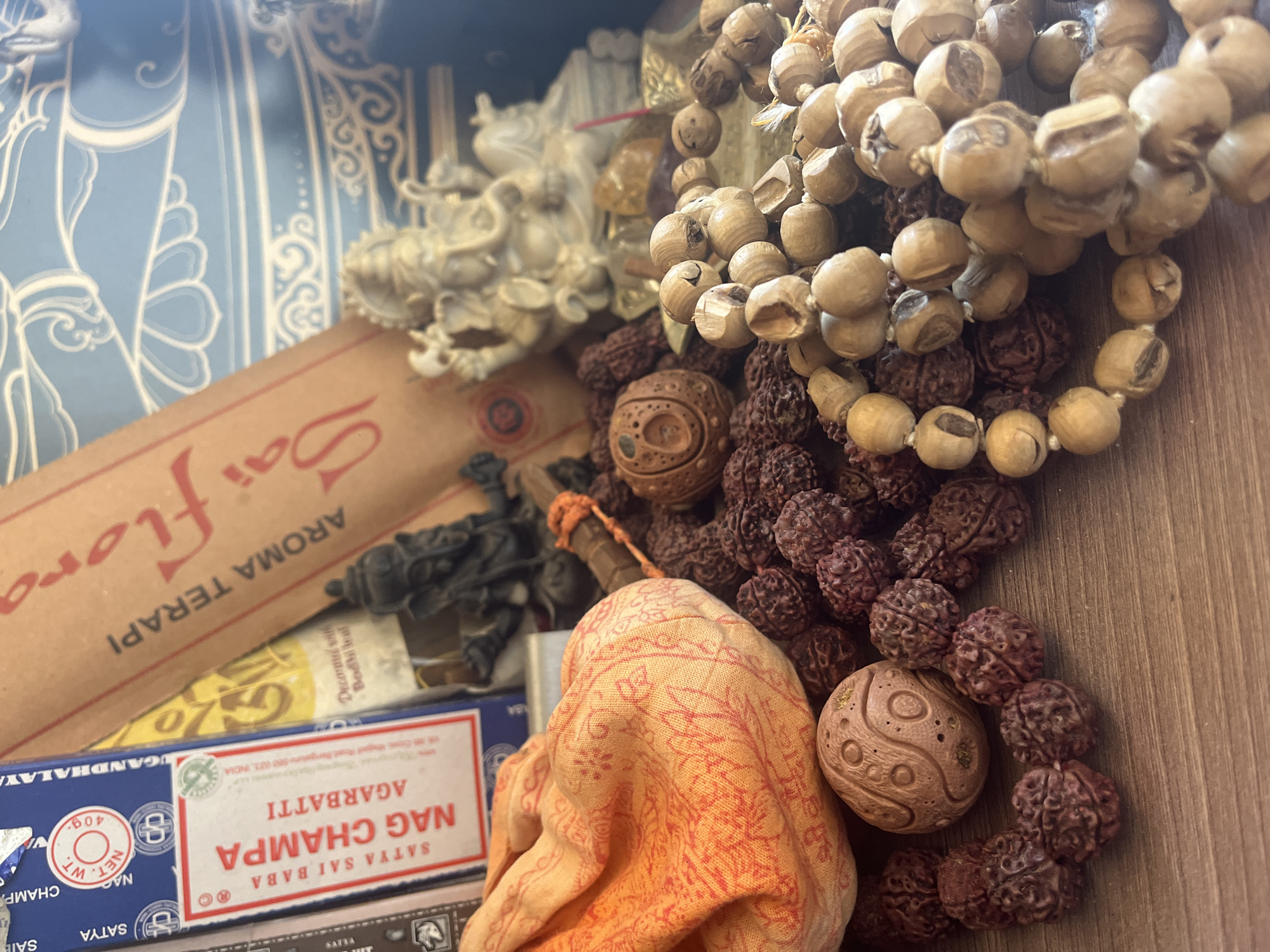 A close-up, top-down view of religious and aromatic items is presented. In the foreground, several strings of beads, varying in color from deep red to light brown, are scattered across a wooden surface. Two ornate, round, carved objects, possibly wooden amulets or prayer beads, are also visible among the strings. A folded orange cloth with a faint red pattern lies to the left. In the background, a cardboard box of "NAG CHAMPA AGARBATTRI" incense sticks is partially visible, along with another box with red lettering and a light yellow design. Behind these, two statuettes are present: a dark, detailed figure of a deity, possibly Shiva, and a lighter, cream-colored statuette of Ganesh. A blue, patterned fabric or wallpaper serves as the backdrop. The scene is well-lit, with soft shadows indicating a gentle light source. There are no people present. The image does not contain any information that would allow for the determination of its location in Balaklavsky District, Ukraine.