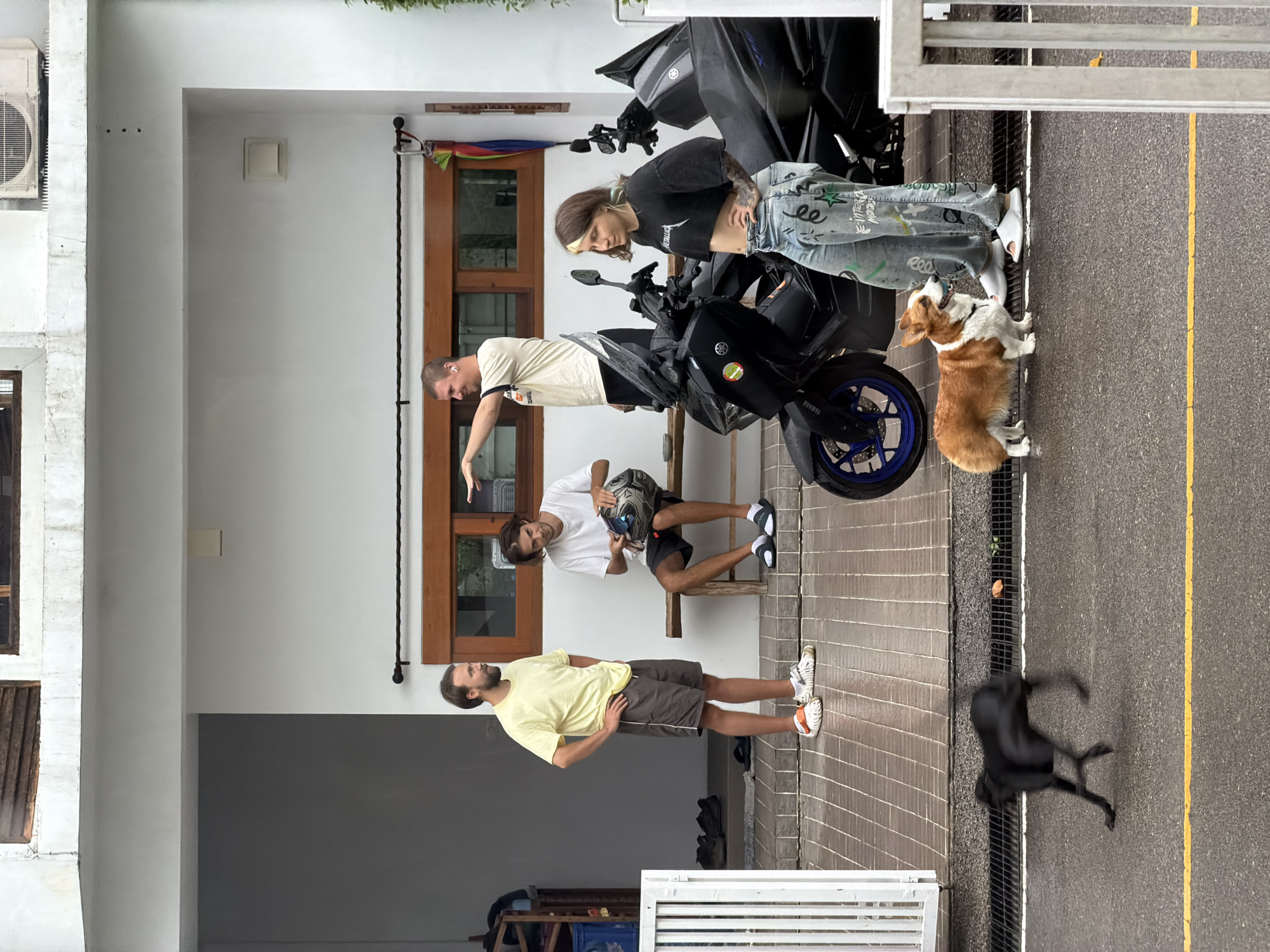 In Chang Phueak, Thailand, a group of four individuals and two dogs are present on a covered porch area adjacent to a building with white walls and wooden accents. On the right, a person with a black t-shirt and light-colored, graffiti-adorned pants stands next to a black motorcycle. To their left, another person in a white t-shirt sits on a wooden bench holding a motorcycle helmet. Further left, a person in a yellow t-shirt and grey shorts stands with their hands on their hips. In the background, a third person in a light-colored t-shirt and black shorts is visible, with their arm extended. In the foreground, on the wet asphalt street, a black dog is captured in motion, and a corgi stands near the motorcycle. The environment appears to be a residential or commercial area with an overhead structure providing shelter.
